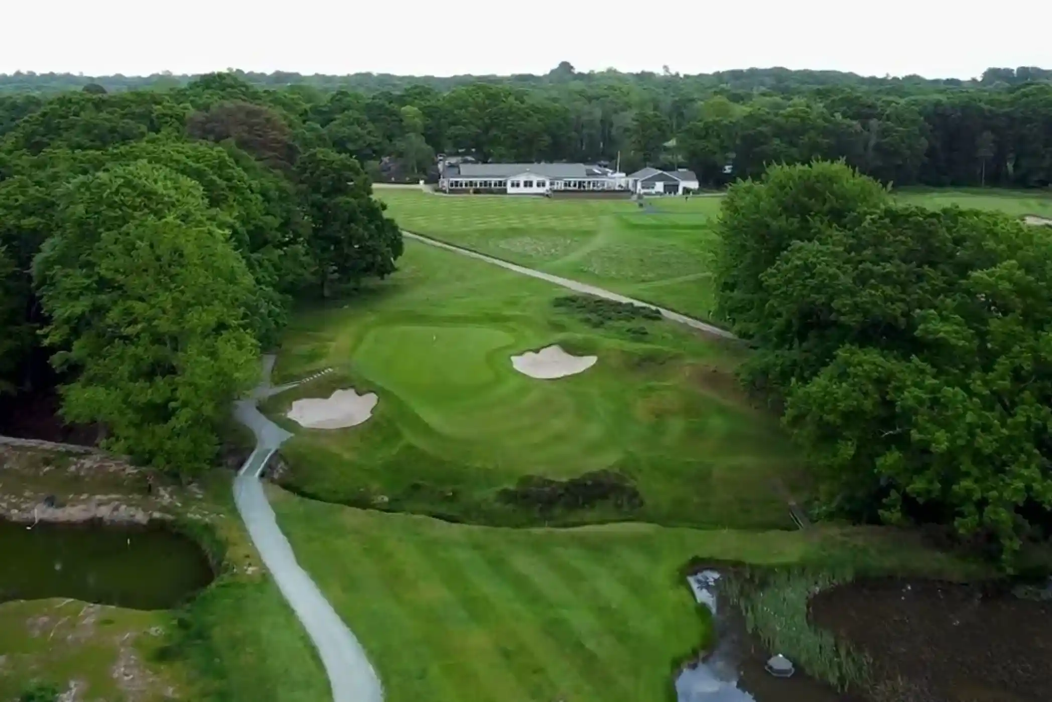 Aerial view of a golf course featuring manicured greens, sand bunkers, and a serene path winding through lush trees. In the background, a clubhouse is visible among the greenery.
