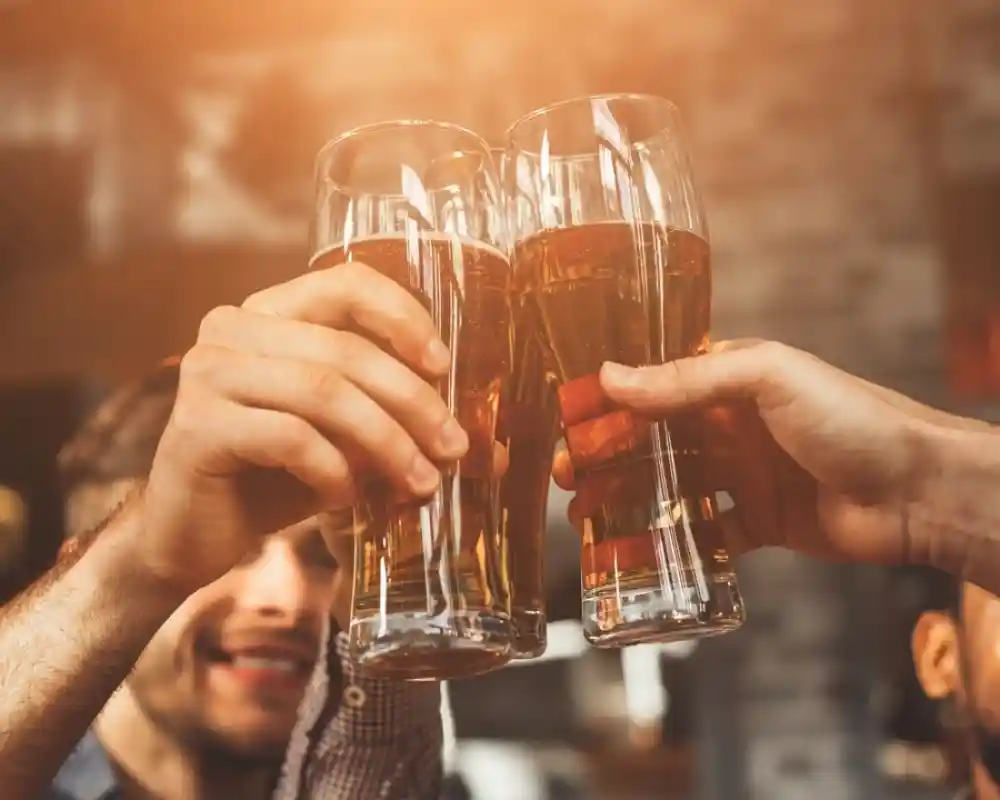 Four glasses of beer clink together in a toast, with smiling people in the background celebrating. Warm light creates a festive atmosphere.