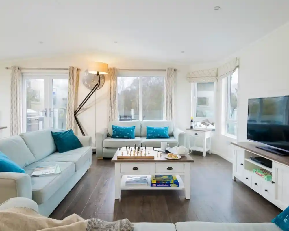 A modern, cozy living room features light blue sofas with decorative pillows, a wooden coffee table with a chess set and snacks, and a large TV. Large windows provide natural light, and there are white curtains and a stylish floor lamp. The floor is dark wood, contributing to the warm ambiance.
