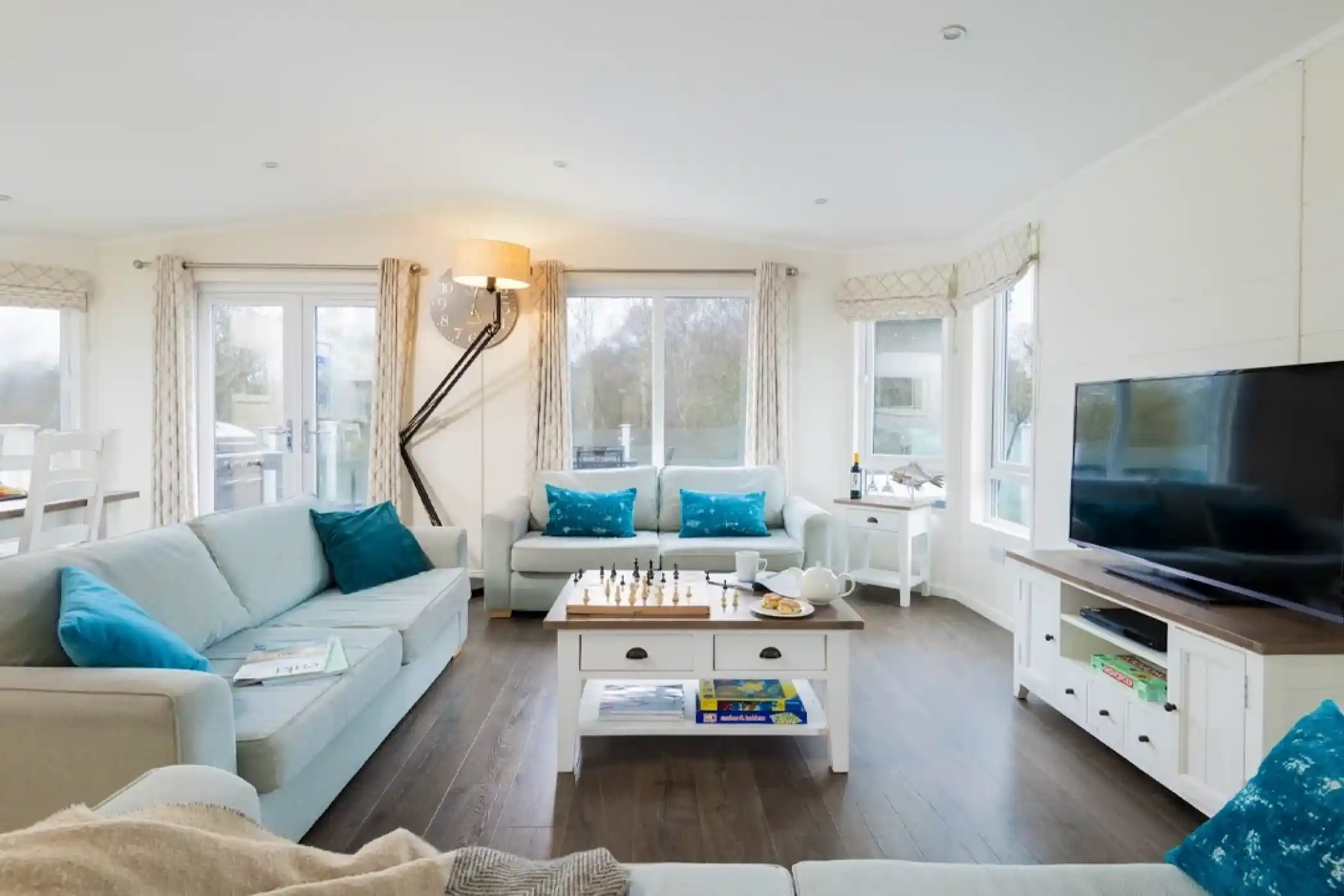 A modern, cozy living room features light blue sofas with decorative pillows, a wooden coffee table with a chess set and snacks, and a large TV. Large windows provide natural light, and there are white curtains and a stylish floor lamp. The floor is dark wood, contributing to the warm ambiance.