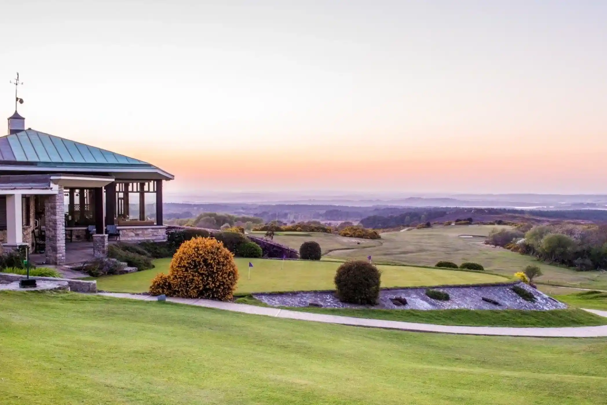 A scenic view of a golf course at sunset, featuring rolling green hills and a colorful sky. In the foreground, a well-manicured lawn leads to a club building with large windows, and a vibrant bush adds a splash of color. The horizon shows distant trees and hills.