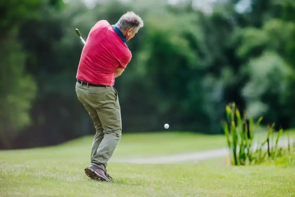 A man in a pink shirt and khaki pants swings a golf club on a lush green golf course. A small white golf ball is airborne, heading towards the distance.