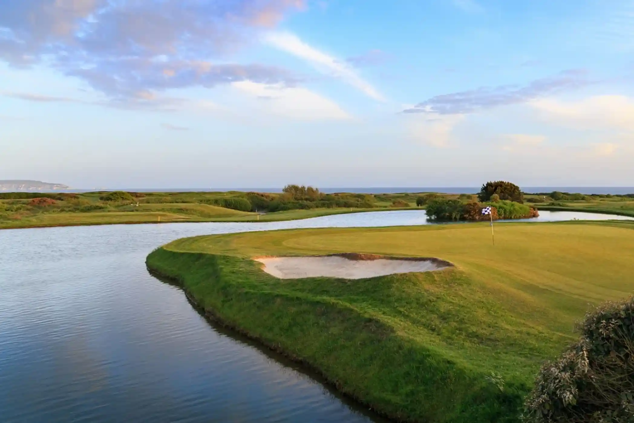 A serene golf course landscape featuring a green fairway, a sand trap, and a water hazard under a clear blue sky with scattered clouds. The coastline is visible in the background.