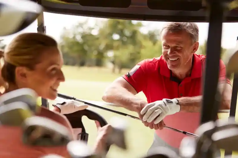 A man in a red polo shirt smiles at a woman sitting in a golf cart, both engaged in a friendly conversation. Golf clubs are visible behind them in the cart, and a green golf course is in the background.