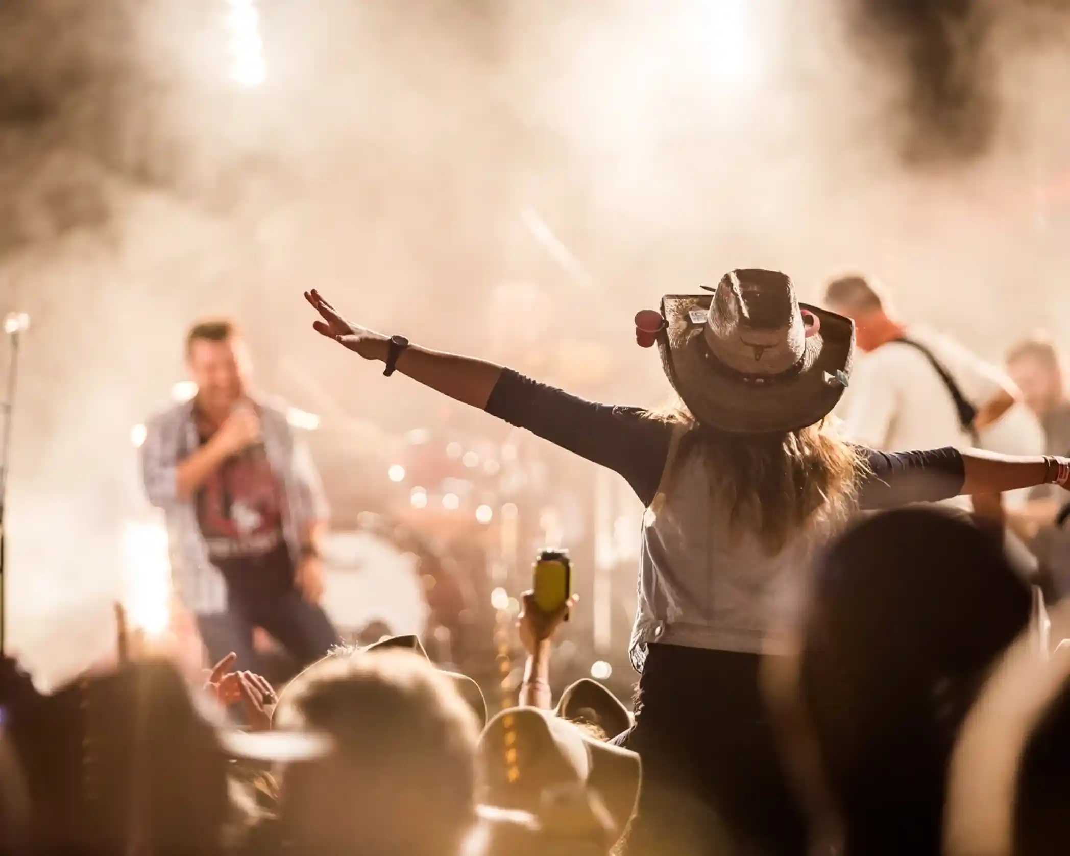 A lady wearing a cowboy hat is pictured on a person's shoulders with her arms spread out wide amongst a crowd of people watching a country music band perform on a stage.