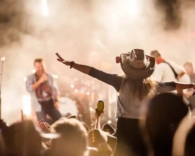 A lady wearing a cowboy hat is pictured on a person's shoulders with her arms spread out wide amongst a crowd of people watching a country music band perform on a stage.
