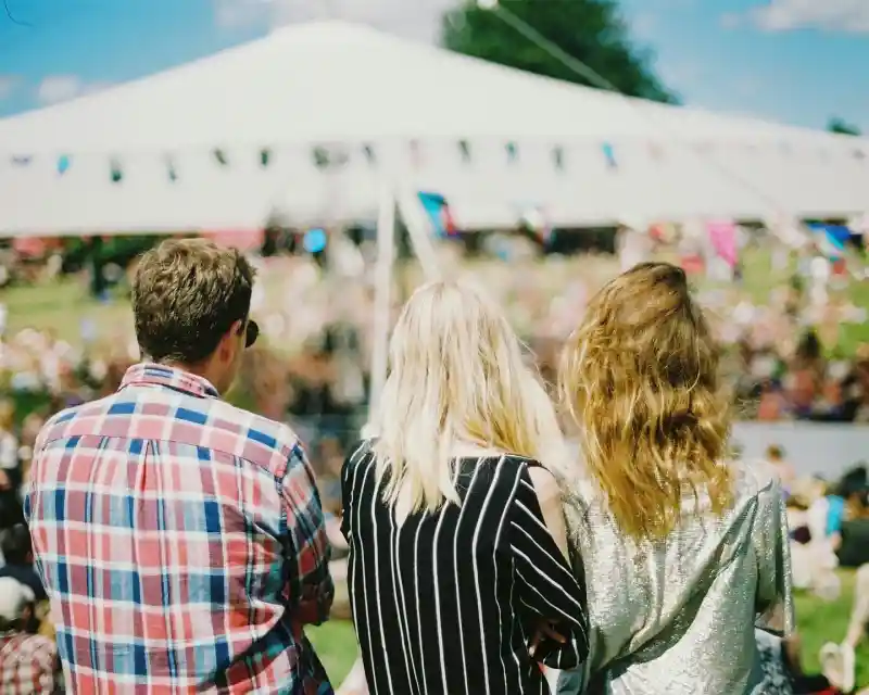 Three people stand with their backs to the camera, watching a festival. A large tent and a crowd are visible in the background, with blue skies overhead. The first person wears a plaid shirt, the second has long blonde hair with a striped top, and the third has wavy hair and a shiny silver top.