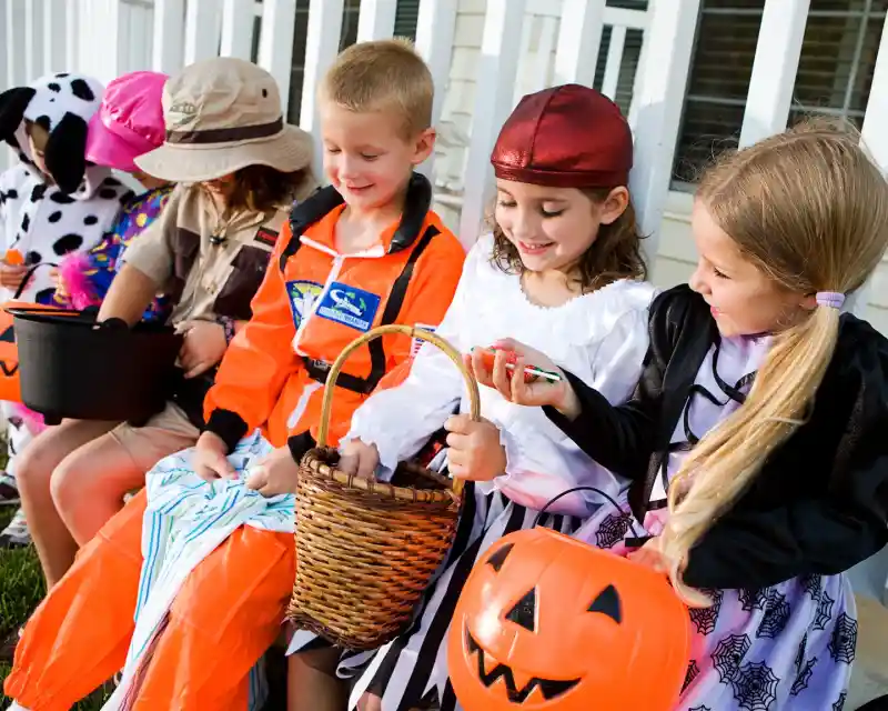 A group of children in Halloween costumes sits on a porch, holding colorful treats and baskets. They include a pirate girl, a child dressed as a cowboy, and others in fun outfits, all smiling and enjoying the festive atmosphere.