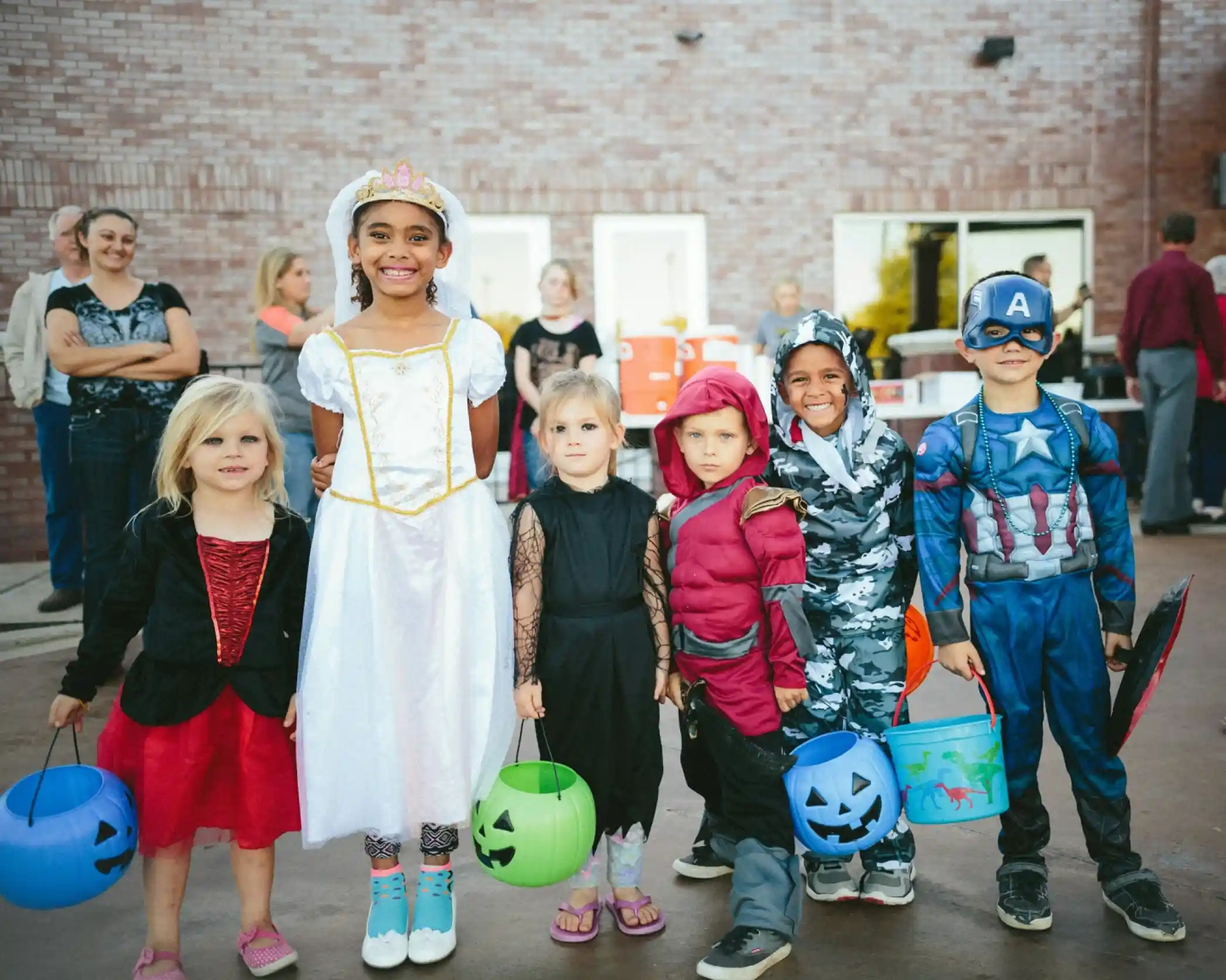 A group of six children in costumes stands together, smiling for the camera. The first child wears a black dress with a red cape, the second is dressed as a princess in a white gown with a tiara, followed by a girl in a black outfit. The next child is dressed as a superhero in a gray camouflage outfit, then a child in a red and black outfit, and finally, a child in a Captain America costume. Each child holds a candy bucket. In the background, adults are seen socializing.