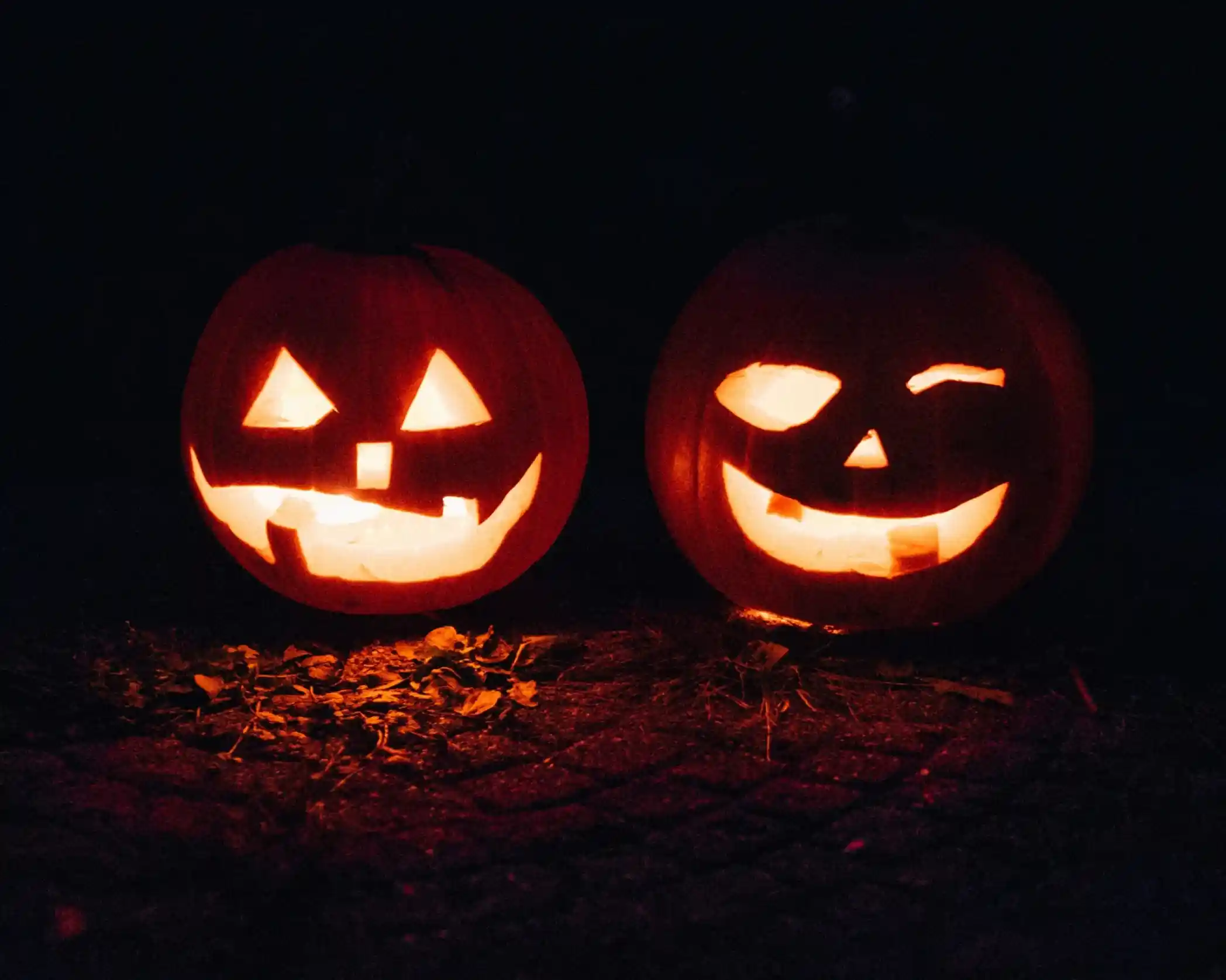 Two carved pumpkins glow in the darkness, one with a classic grinning face and the other with a playful wink and smile. Both are surrounded by fallen leaves on the ground.