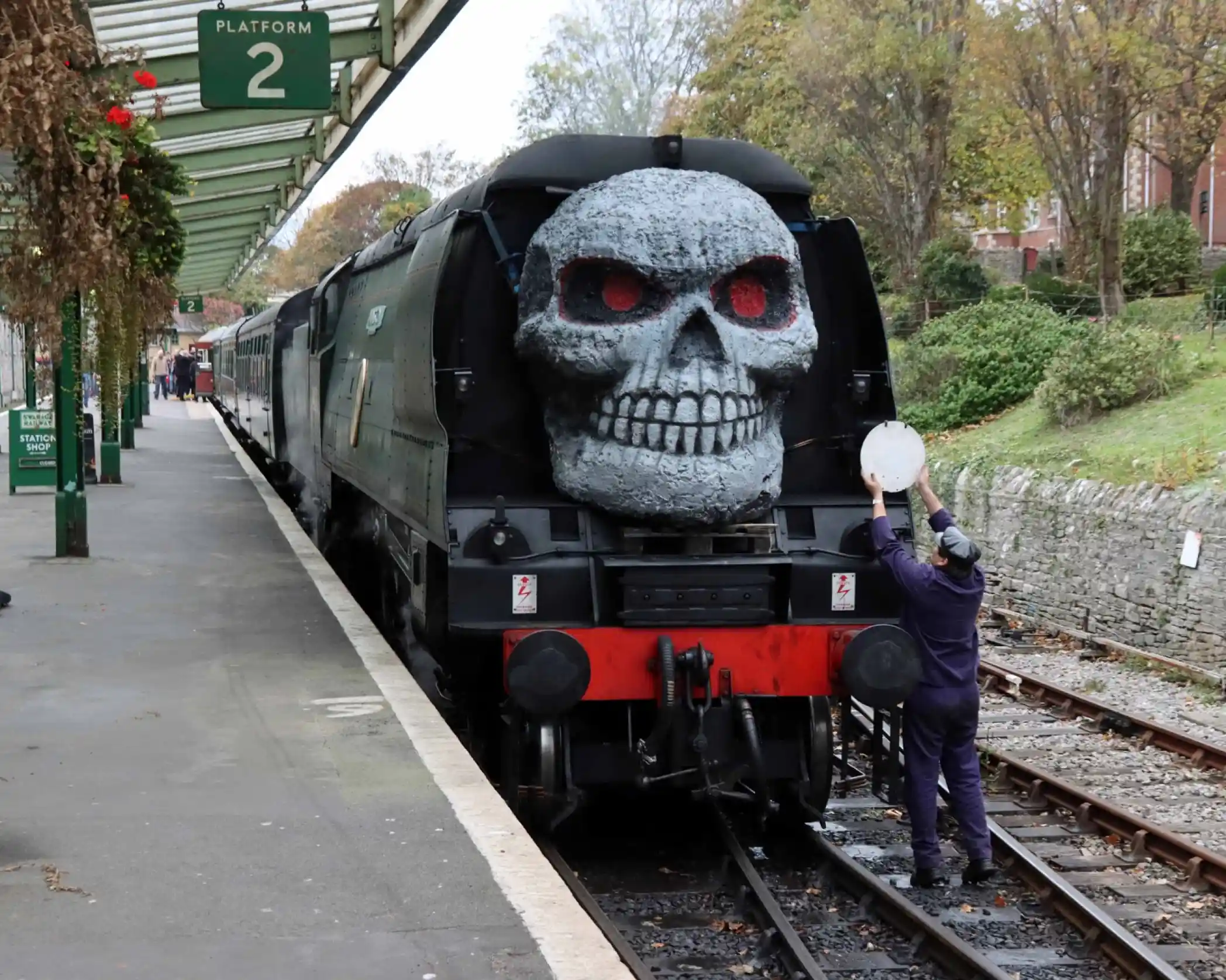 A steam train with a large skull decoration on its front is parked at a platform. A worker in a blue uniform stands nearby, holding a circular object. Surrounding trees and a brick wall are visible in the background.