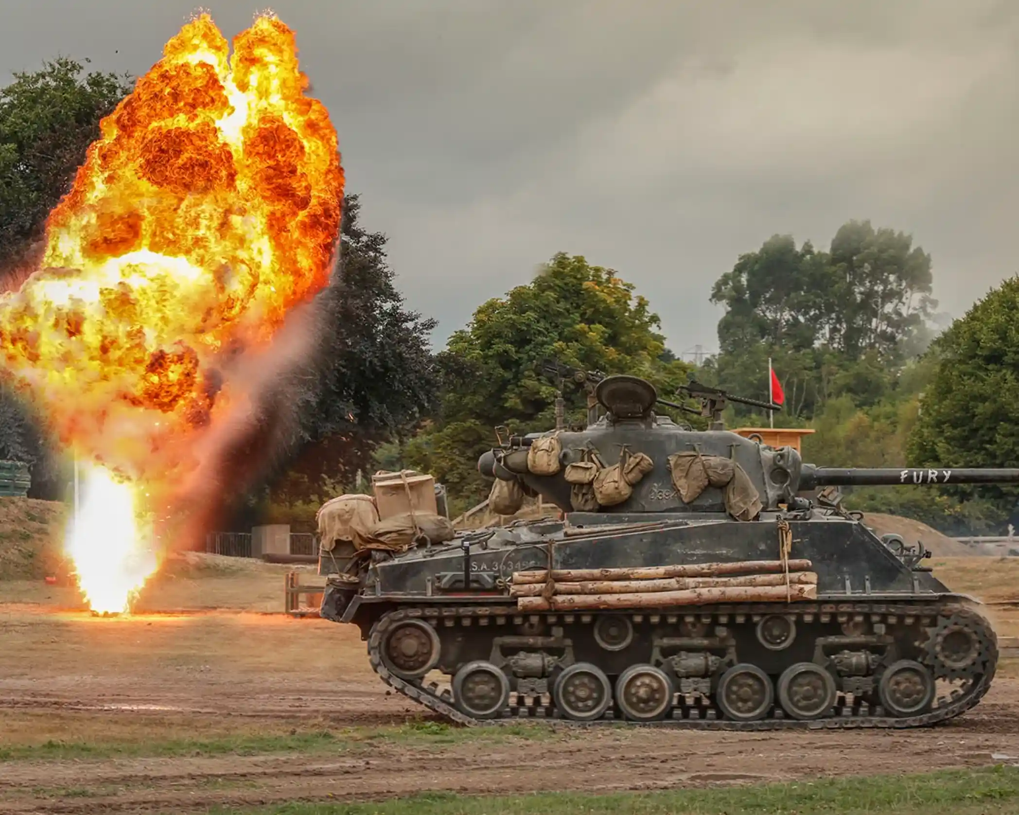 A vintage tank labeled "Fury" stands on a grassy field with a large explosion of fire and smoke erupting nearby. Trees and a cloudy sky are visible in the background.