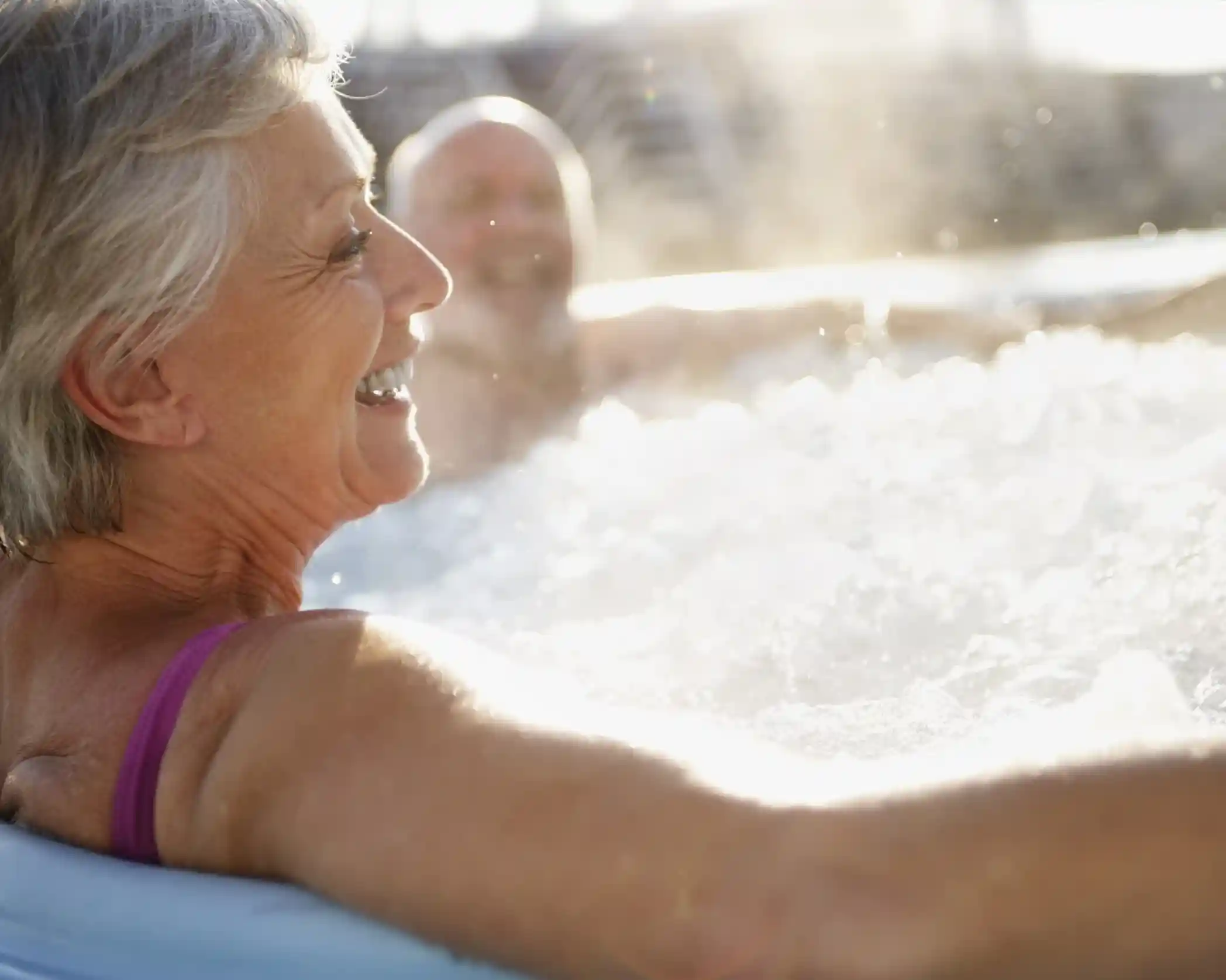 A woman with gray hair smiles while relaxing in a hot tub, with steam rising in the air. A man can be seen in the background, also enjoying the warm water.
