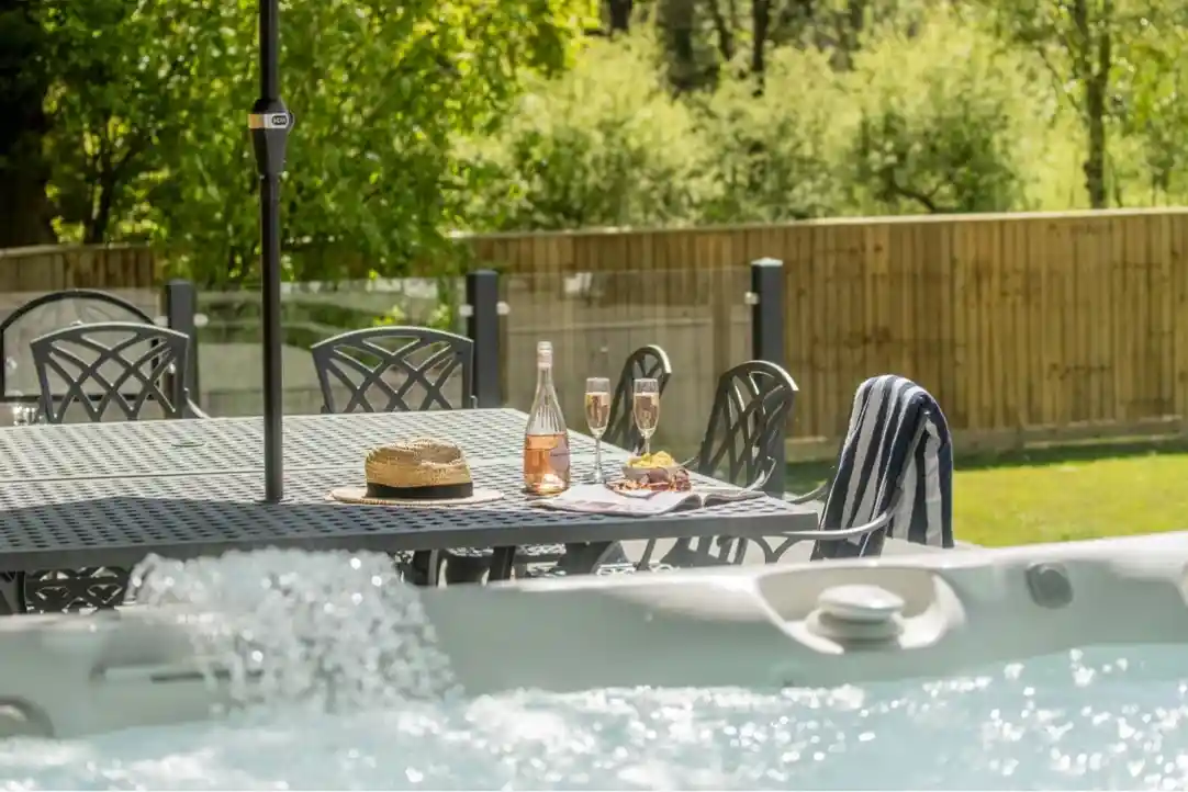 A sunny outdoor scene featuring a hot tub in the foreground, accompanied by a table set with a bottle of rosé, two glasses, a straw hat, and a striped towel. Lush greenery and a wooden fence are visible in the background.