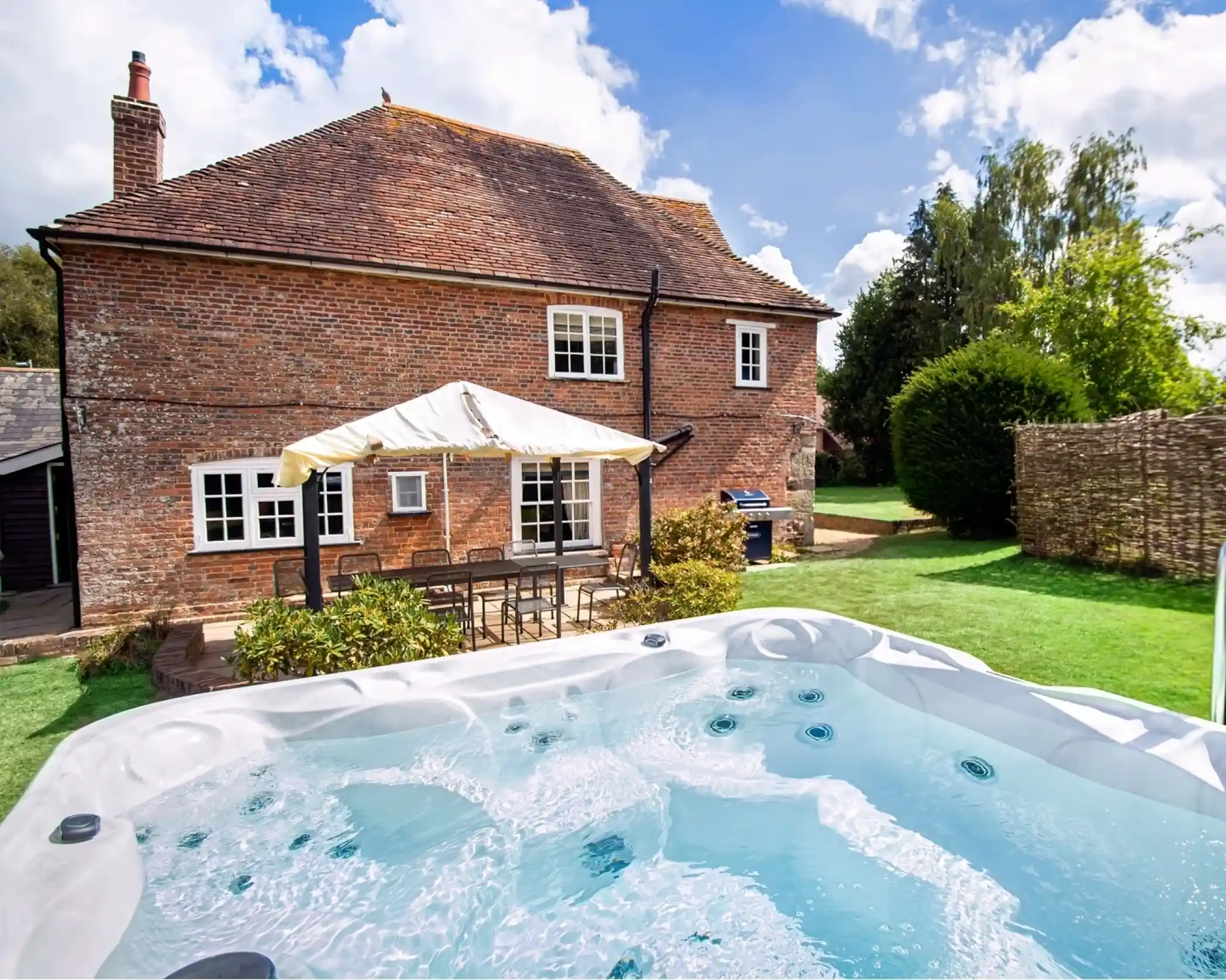 A spacious backyard with a hot tub in the foreground, a brick house with white window frames in the background, and a well-maintained garden under a partly cloudy sky.