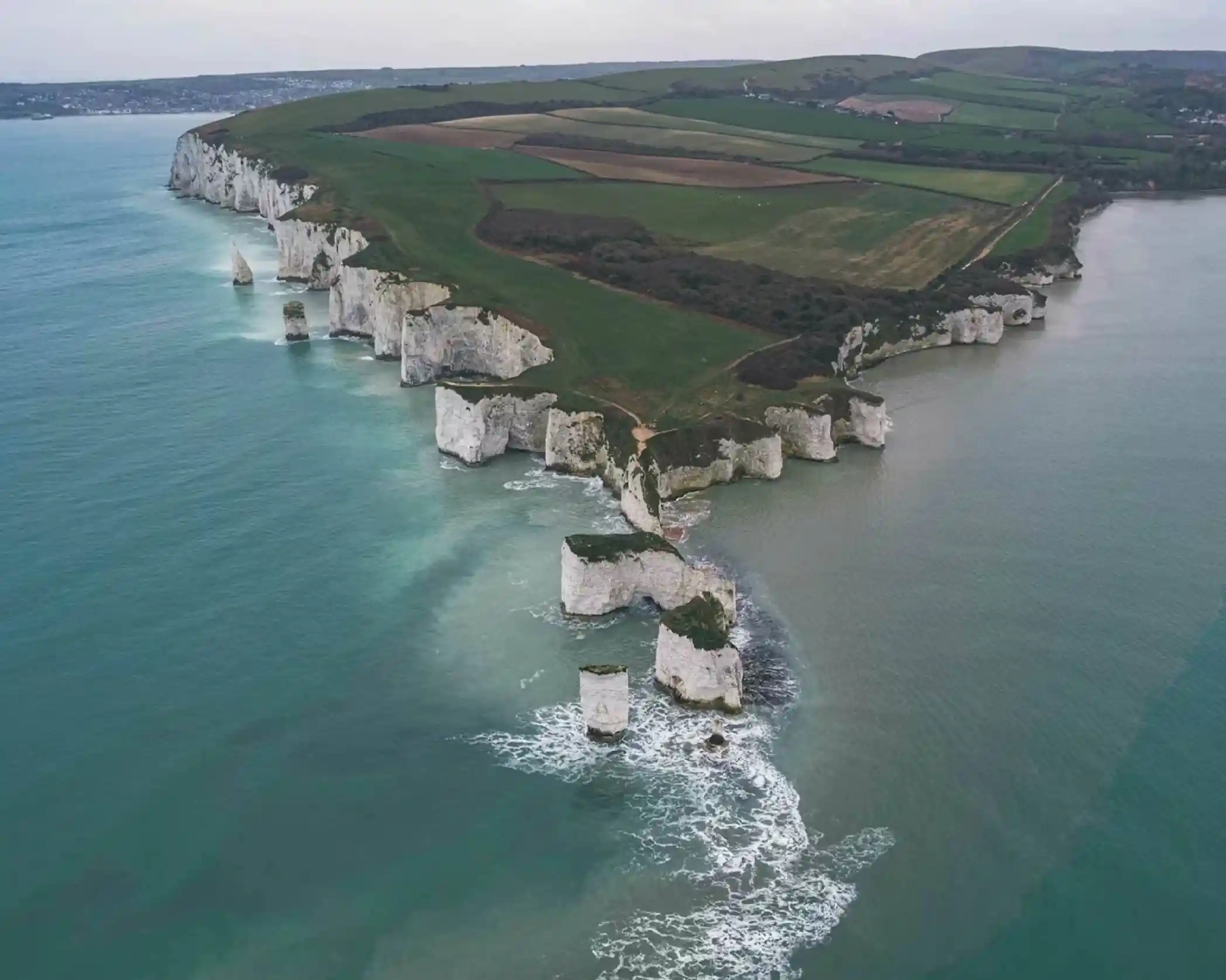 Aerial view of rugged white cliffs meeting a turquoise sea, with green hills and farmland in the background. The cliffs feature distinct rock formations and gentle waves splashing against them.