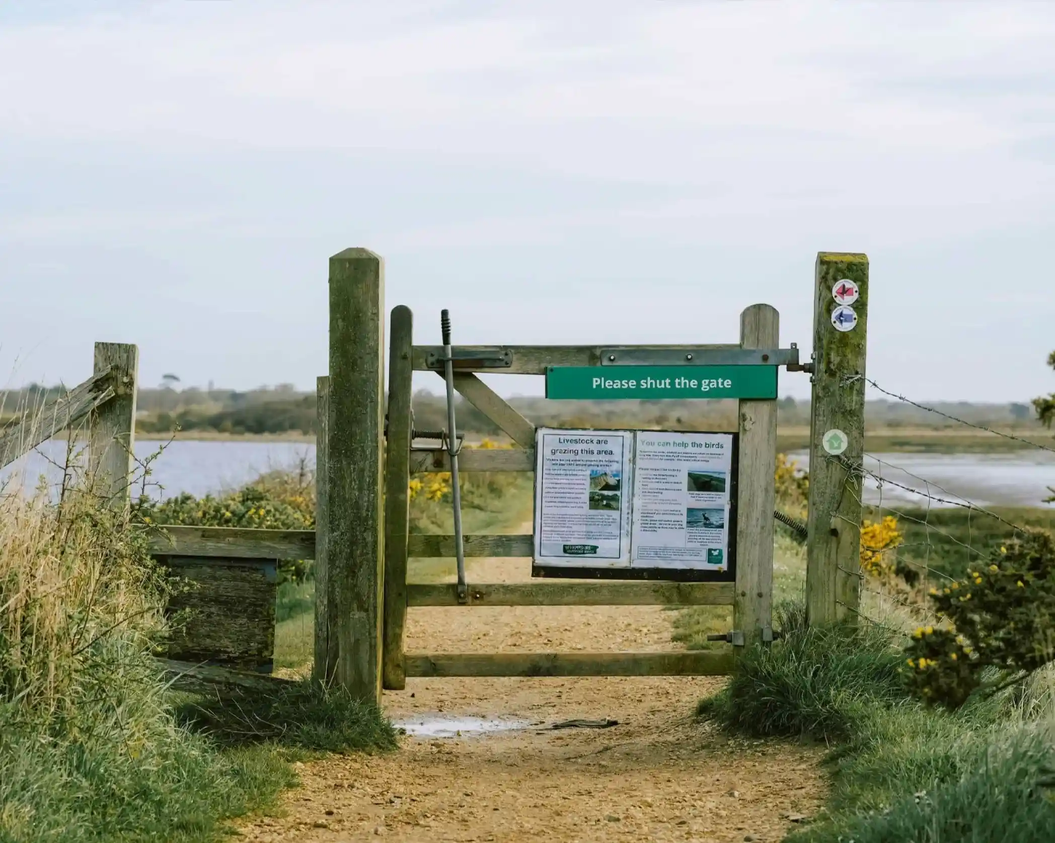 A wooden gate with a sign that says "Please shut the gate" opens to a path leading towards a scenic landscape with grass and water in the background. Informational panels are attached to the gate.