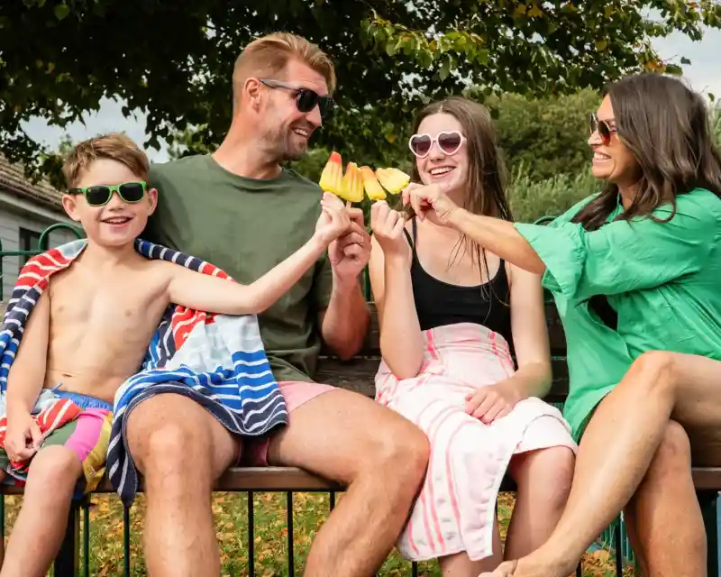 A family of four enjoys a sunny day outdoors, sitting on a bench. They are wearing sunglasses and beach attire, smiling and holding colorful popsicles while playfully toasting them together.