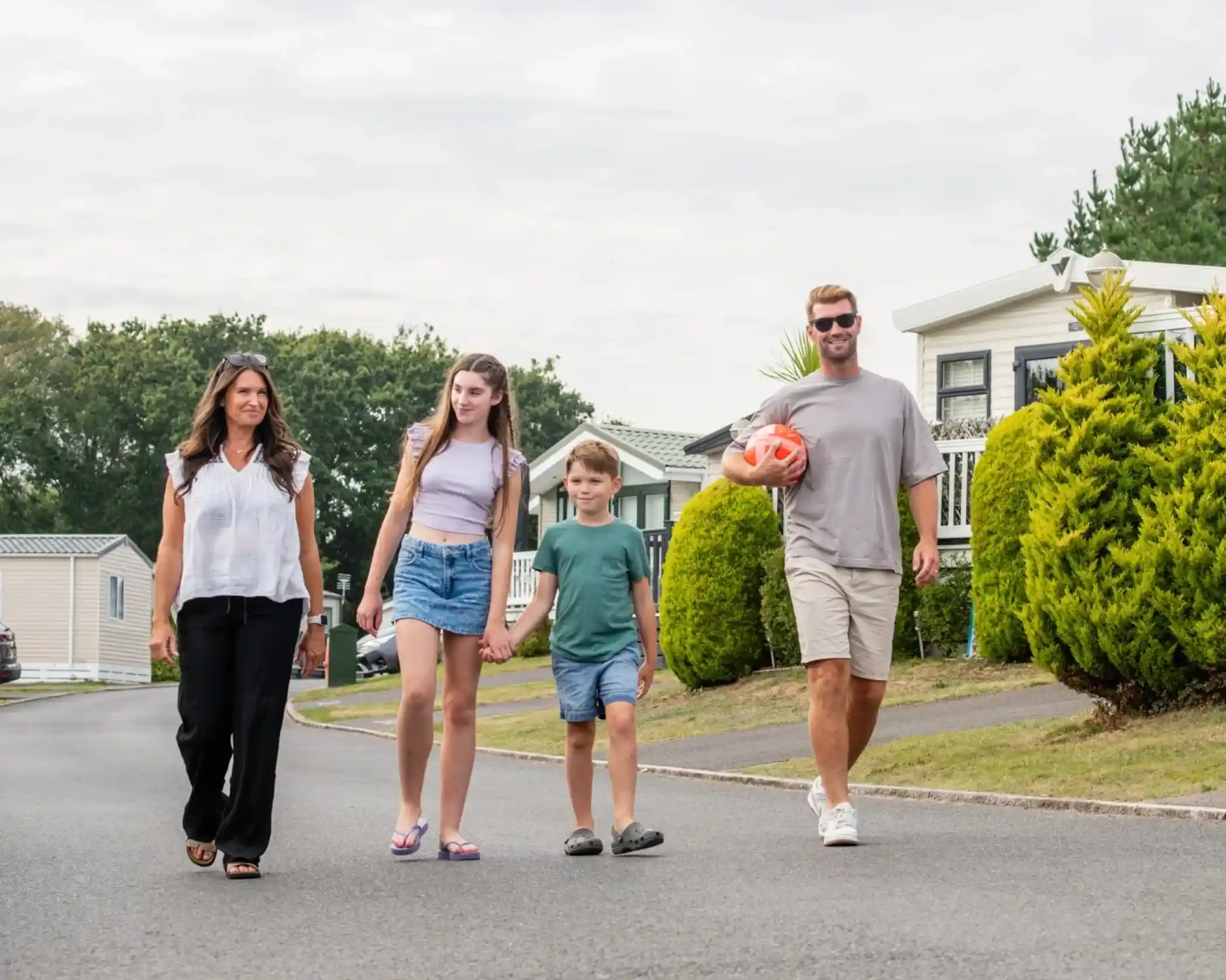 A family walks together along a quiet suburban street, enjoying a leisurely day. Two children hold hands while their parents walk beside them. One parent carries a soccer ball, and they are surrounded by green bushes and homes in the background.