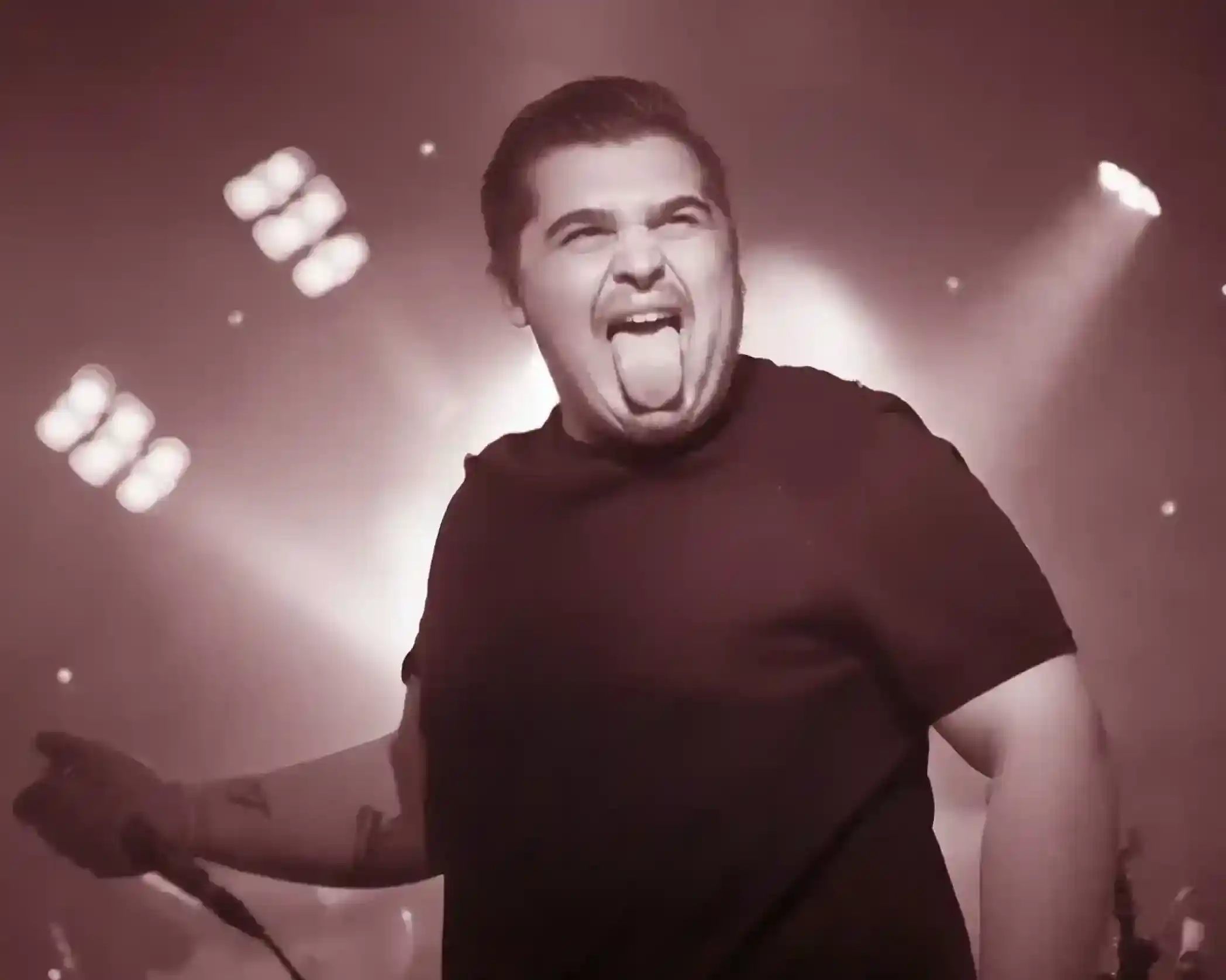 A performer passionately singing on stage, with a wide grin and tongue out, under bright stage lights.