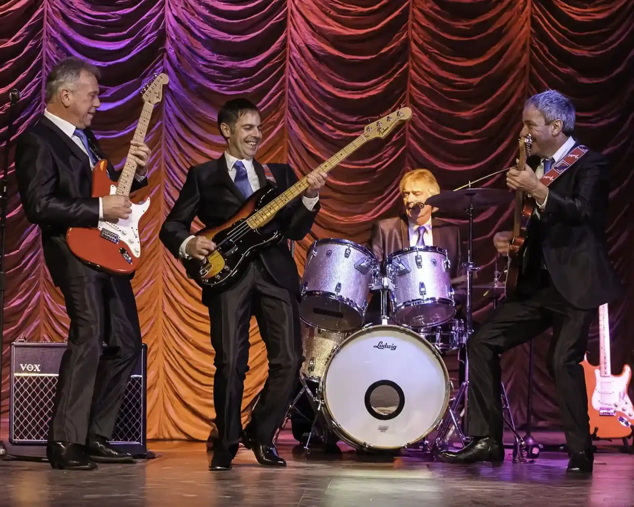 Four musicians in black suits perform on stage. Two are playing electric guitars, one is on bass, and a drummer can be seen in the background. Colorful stage lighting creates an energetic atmosphere.