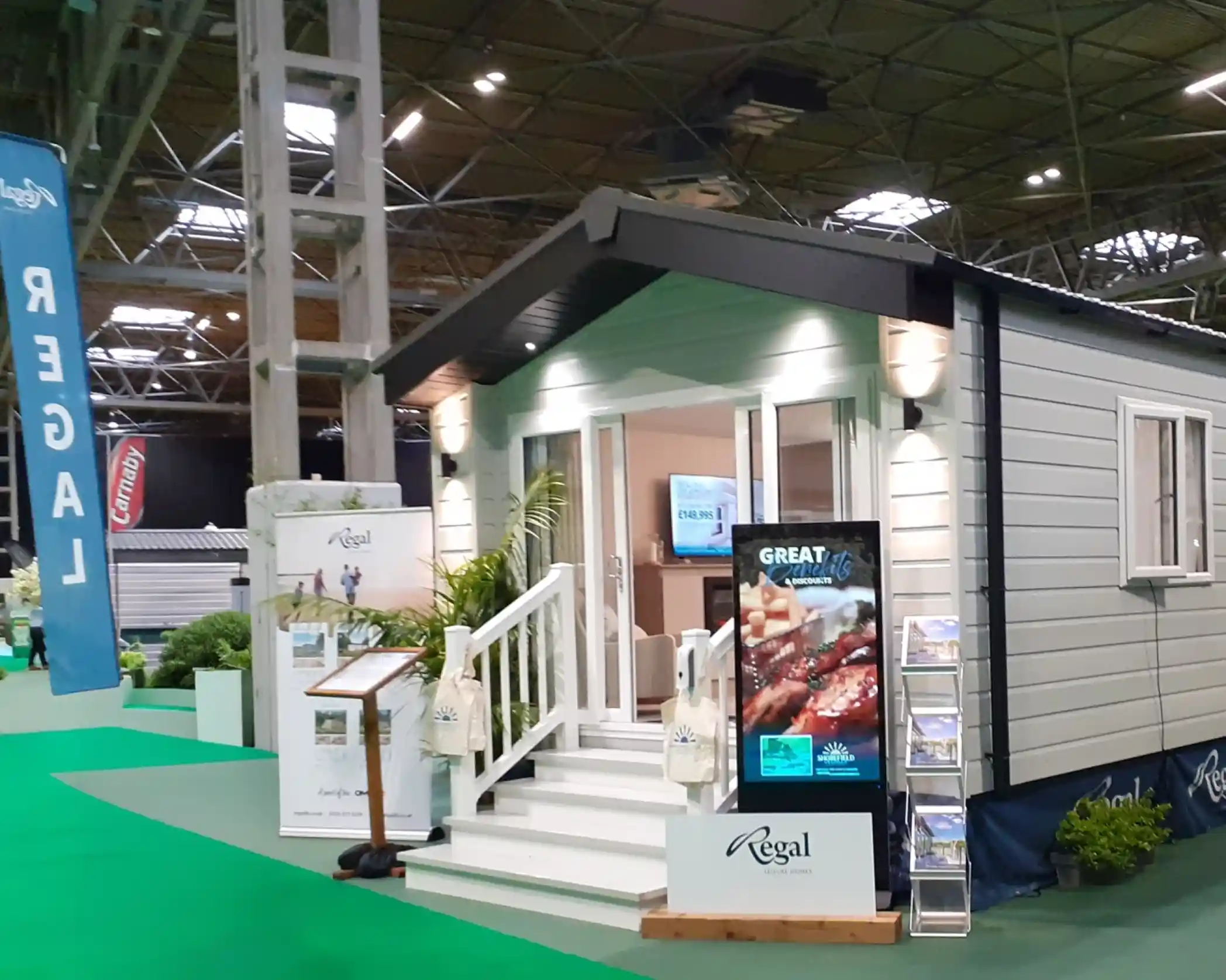 A modern, stylish cabin with a light blue and gray exterior, featuring a front porch with steps. The entrance is adorned with two outdoor lights, and a large digital display showcases promotional content. Green carpet and banners are visible in the exhibition space surrounding the cabin.