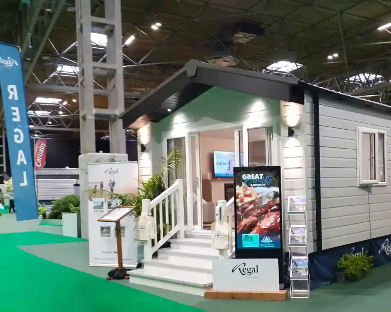 A modern, stylish cabin with a light blue and gray exterior, featuring a front porch with steps. The entrance is adorned with two outdoor lights, and a large digital display showcases promotional content. Green carpet and banners are visible in the exhibition space surrounding the cabin.