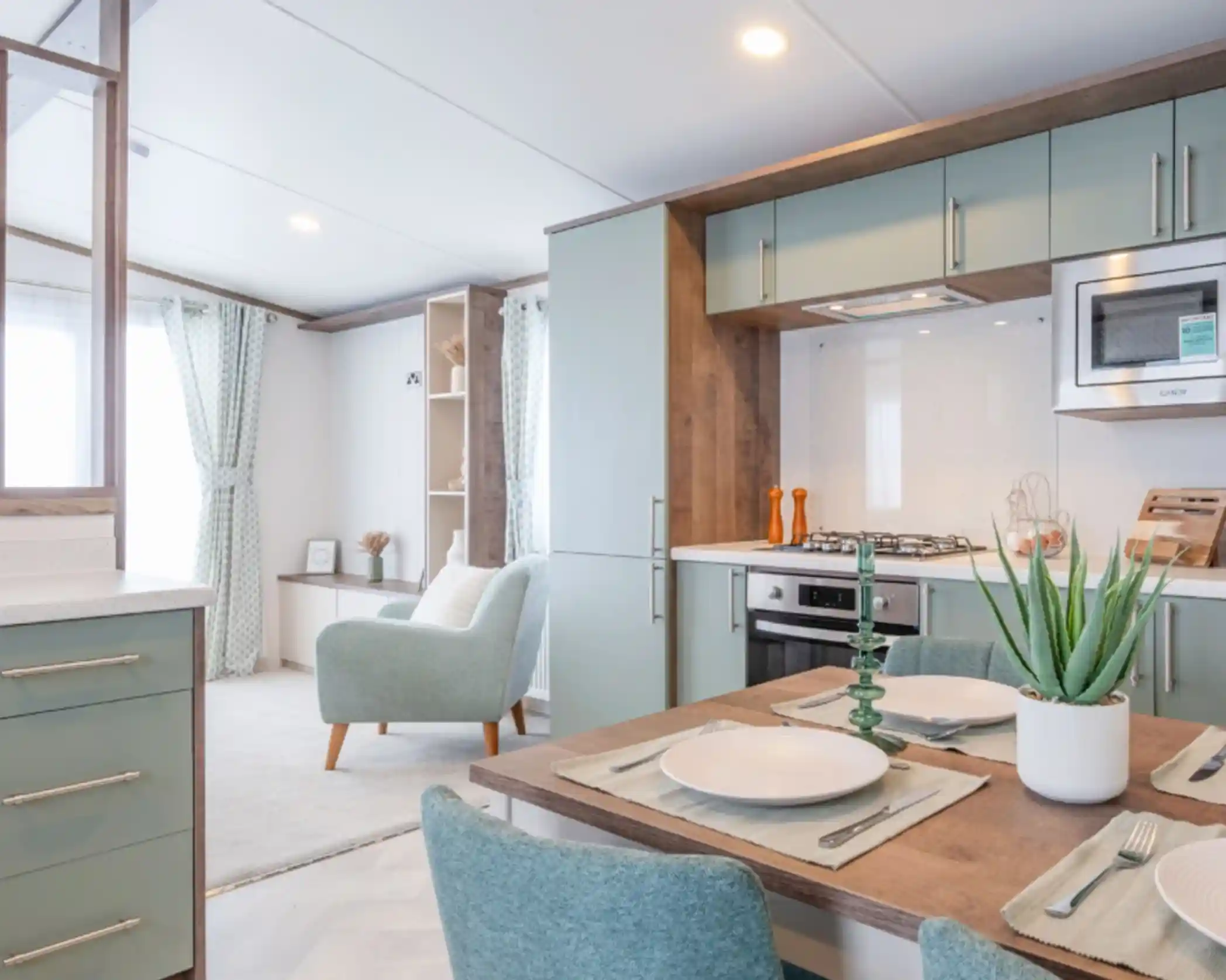 A modern kitchen and dining area featuring mint green cabinetry, a white countertop, and a dining table set for four. A cozy armchair is placed next to a large window with light curtains, and a potted plant adds a touch of greenery.