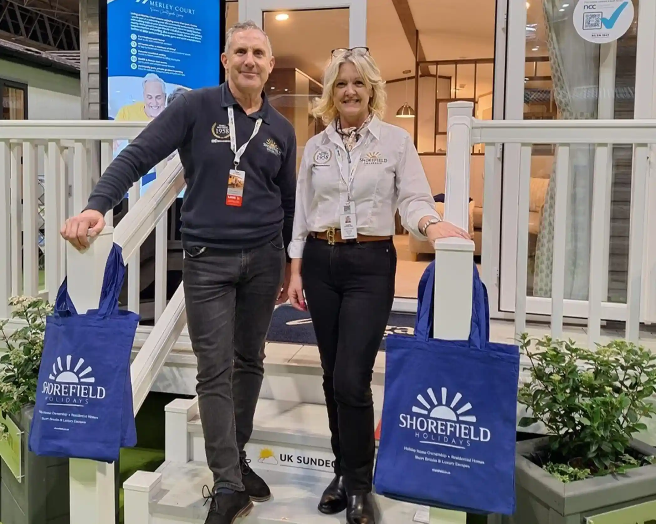 Two individuals stand on the steps of a display booth, smiling and posing for the camera. They are holding blue tote bags featuring the logo of Shorefield Holidays. The background shows a welcoming interior with promotional materials.