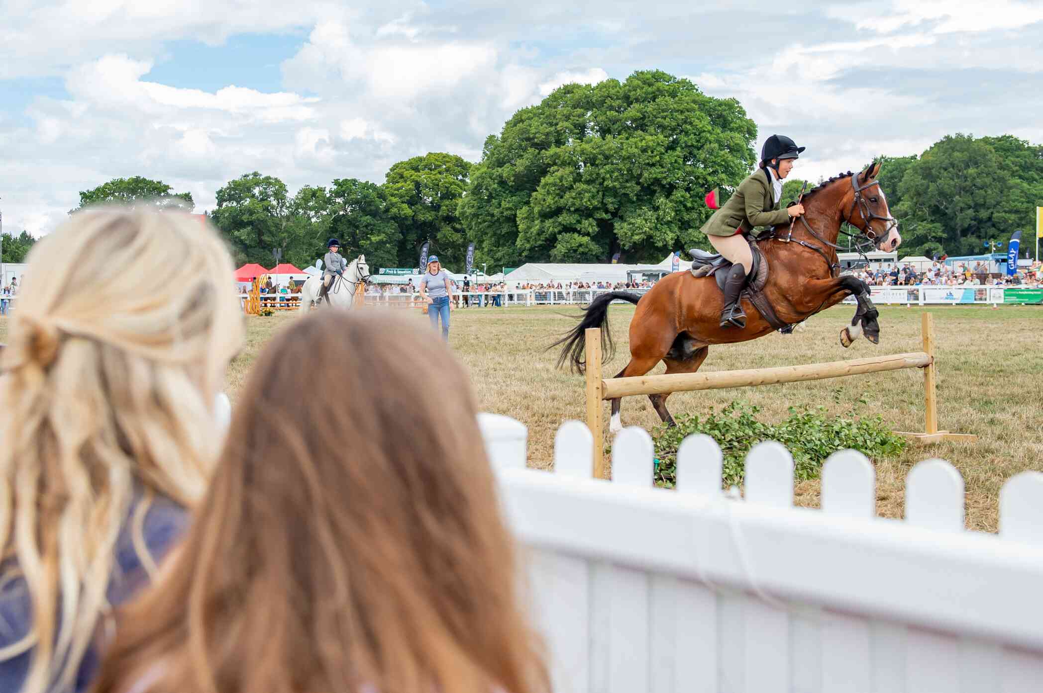A rider in a green jacket and black helmet is jumping over a hurdle on a brown horse during an outdoor equestrian event. In the foreground, two spectators are watching from behind a white fence. Lush trees and a crowd are visible in the background under a partly cloudy sky.
