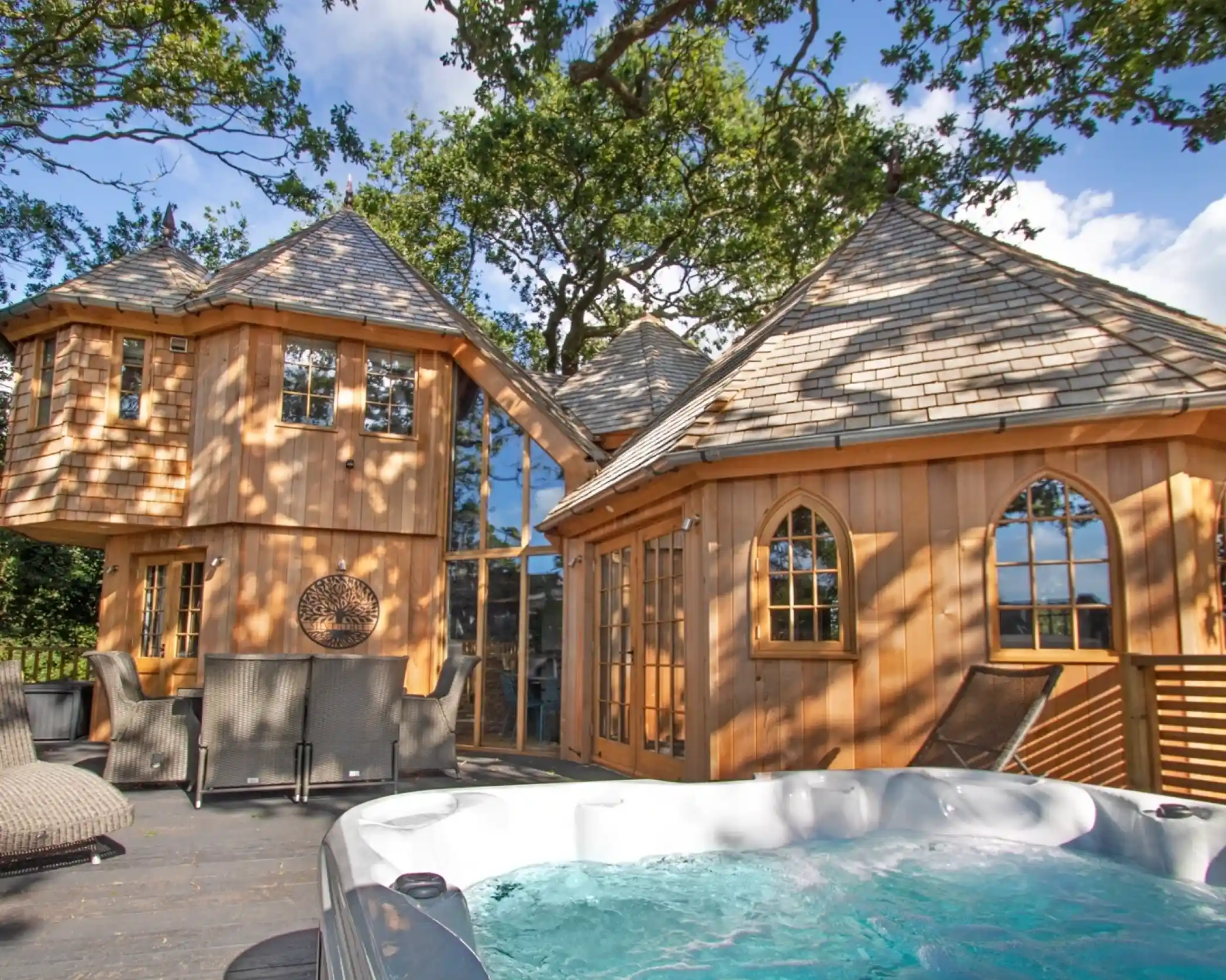 A wooden house with multiple peaks and large windows surrounded by trees. A hot tub sits on the deck in the foreground, with outdoor seating visible nearby. The sky is partly cloudy, creating dappled sunlight on the structure.