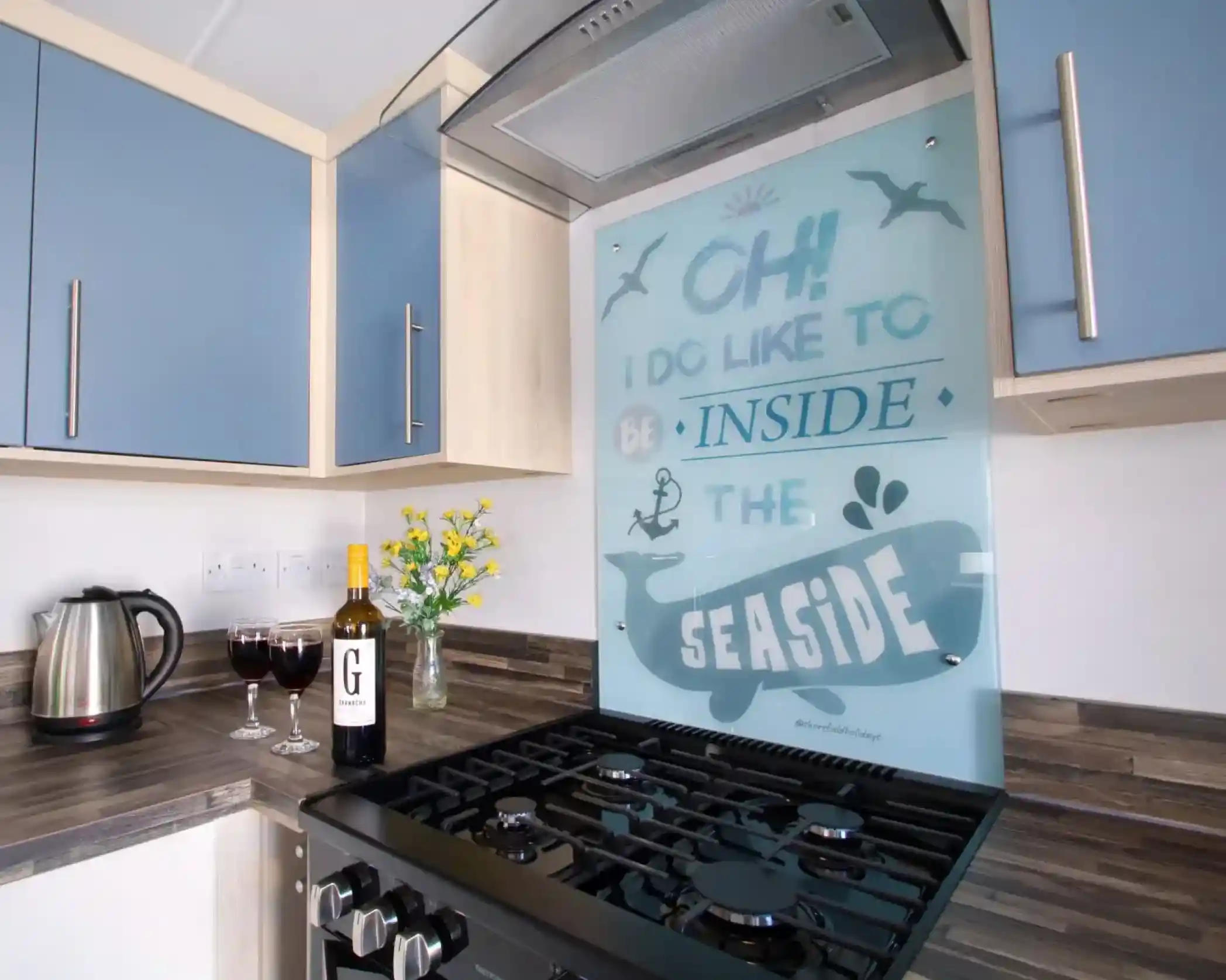 A modern kitchen with blue cabinets and a gas stove. The backsplash features a design that says, "Oh! I do like to be inside the seaside." A kettle, a bottle of wine, two glasses of red wine, and a small vase with yellow flowers are on the countertop.