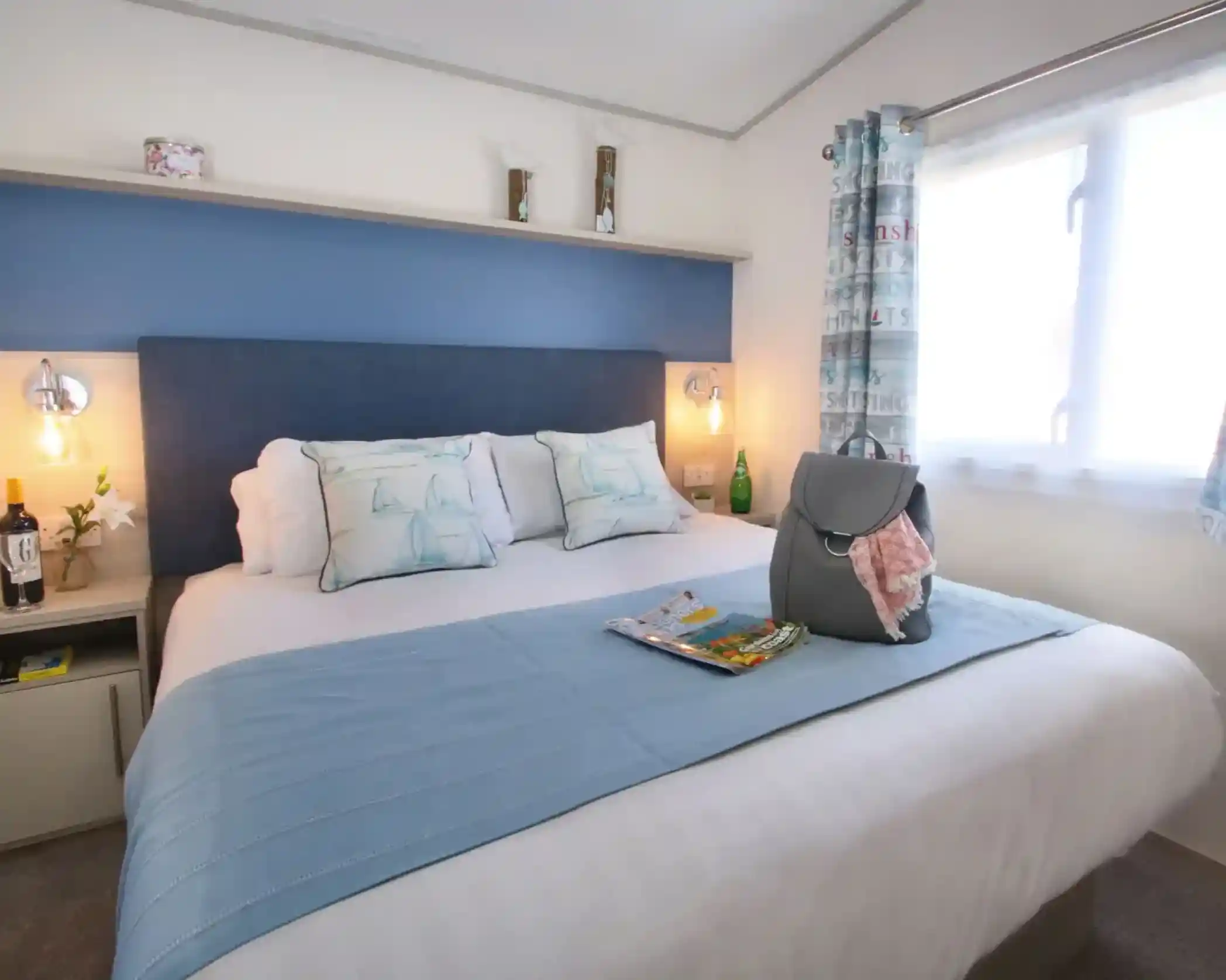 A cozy bedroom featuring a comfortable queen-sized bed with a light blue throw blanket. The headboard is dark blue, and there are two pillows with a nautical design. A small bedside table holds a bottle of water and a magazine. Natural light streams in through a window adorned with sheer curtains. A handbag rests on the bed.