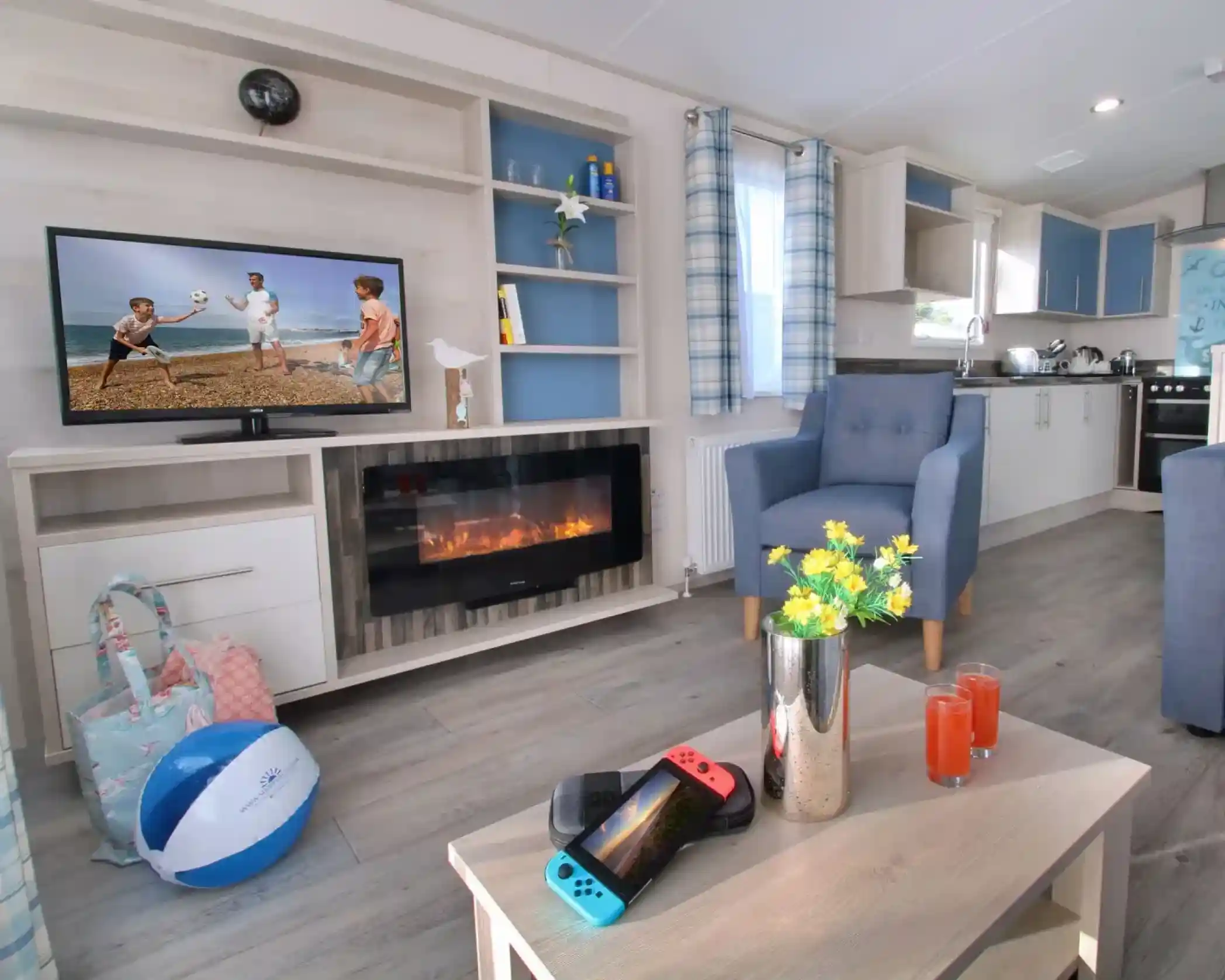 A cozy living area features a modern TV displaying a beach scene, a comfy blue armchair, and a coffee table with two drinks and a Nintendo Switch. A decorative vase with yellow flowers sits atop the table, and beach accessories are scattered around. The kitchen is visible in the background.