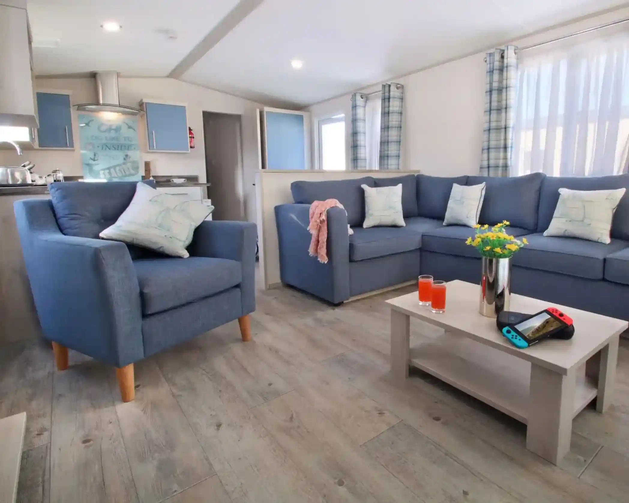 A cozy living room with a light wood floor. There are two blue sofas adorned with patterned cushions and a comfortable blue armchair. A small coffee table holds a vase of flowers, orange drinks, and a Nintendo Switch console. Natural light streams in through large windows with light curtains.