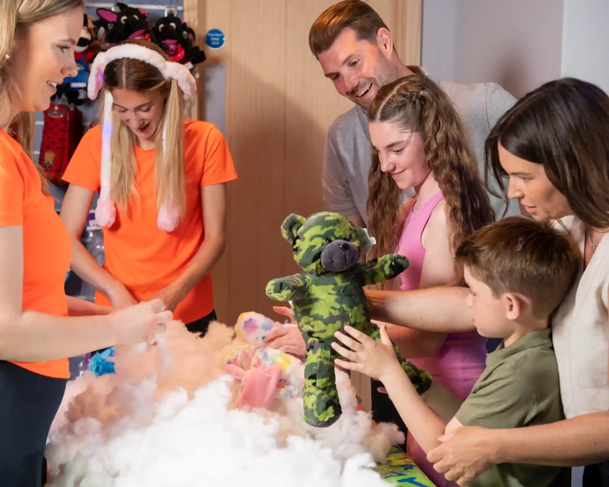 A group of six people gathers around a table covered in fluffy white stuffing and colorful fabric. A child holds up a green camo teddy bear while others look on, smiling and engaged in a creative activity. Two of the adults are helping with the process, and the atmosphere is joyful and playful.