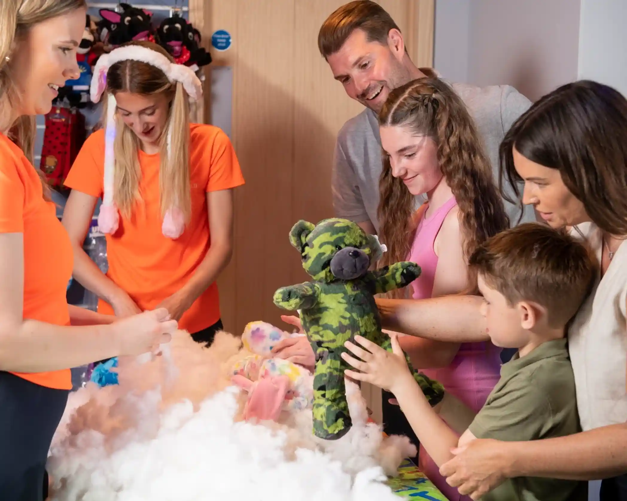 A group of six people gathers around a table covered in fluffy white stuffing and colorful fabric. A child holds up a green camo teddy bear while others look on, smiling and engaged in a creative activity. Two of the adults are helping with the process, and the atmosphere is joyful and playful.
