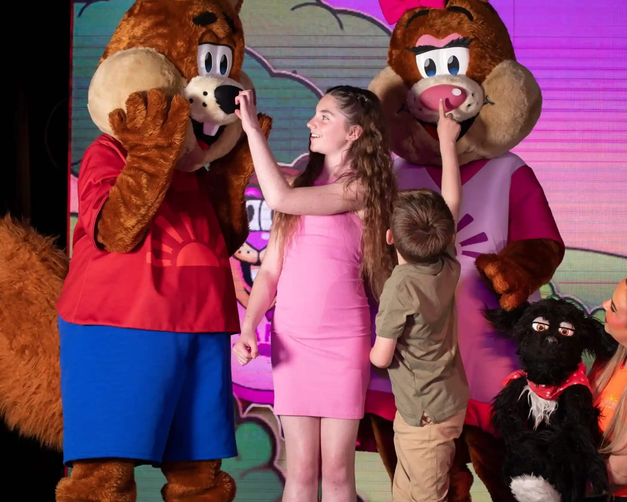 A girl in a pink dress stands between two large animal mascots, who are both wearing shirts. One mascot is playfully touching her nose, while the other mascot has its hands up to its face. A boy in a green shirt is leaning in to touch one mascot as well. A puppet and a character in the background are also visible, creating a cheerful scene.