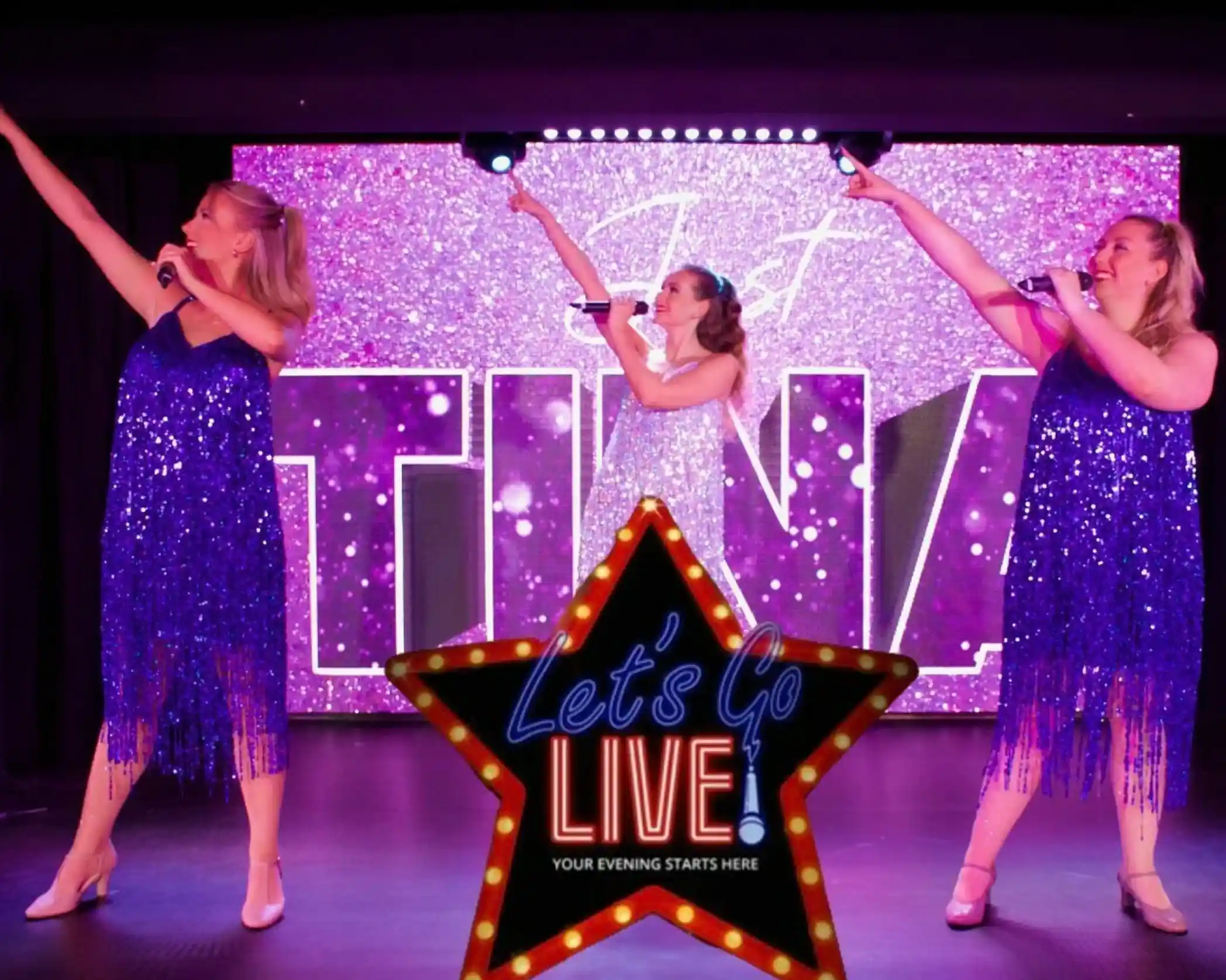 Three female performers in sparkly blue and silver dresses sing on stage, striking a pose with one arm pointing upward. Behind them is a glittery pink backdrop with the word 'TINA' and stage lights. In the foreground, a large illuminated star-shaped sign reads 'Let's Go LIVE - Your Evening Starts Here.