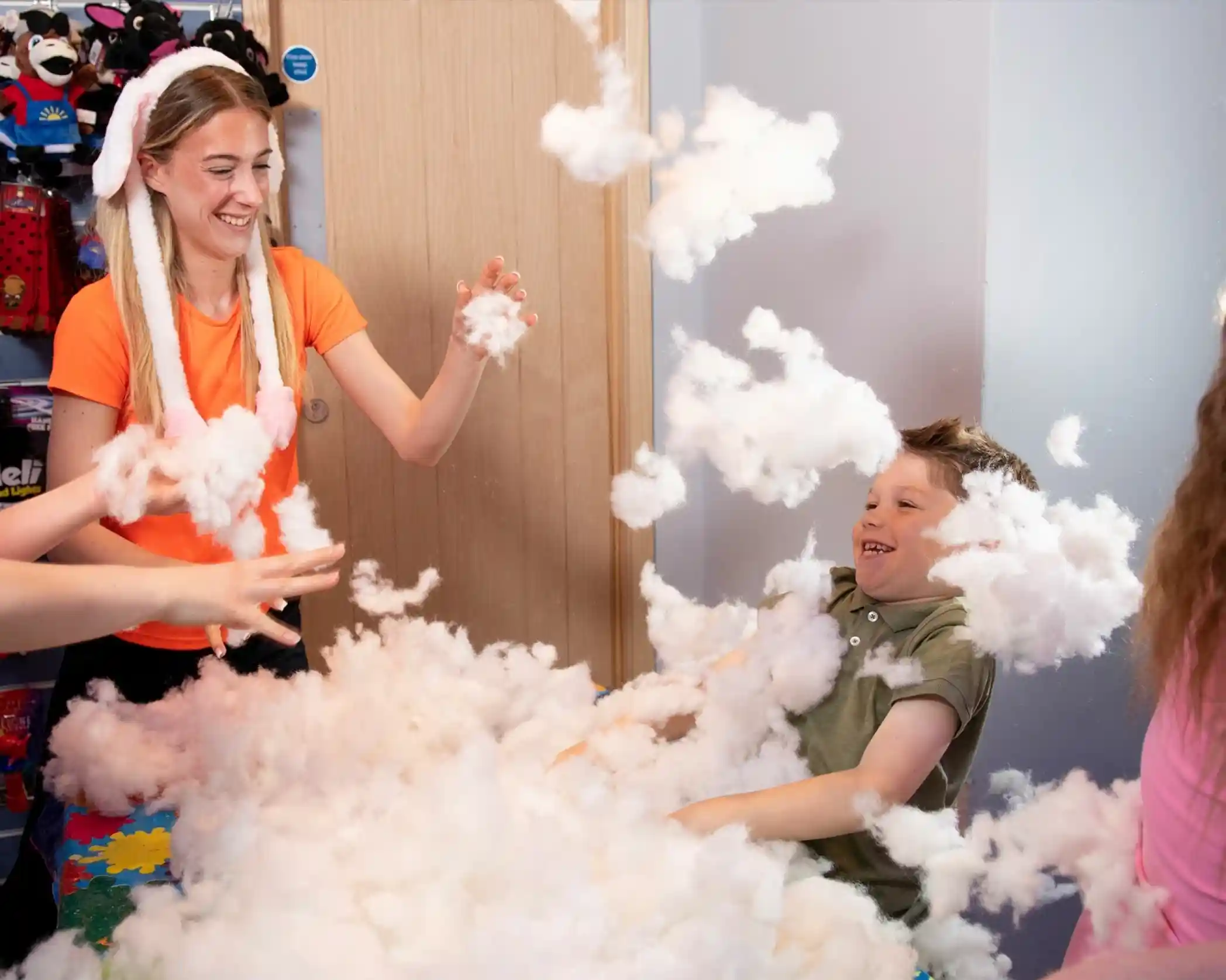 A young woman wearing bunny ears and an orange shirt smiles while playing with synthetic stuffing material alongside a laughing boy and other children. The air is filled with fluffy white stuffing, creating a fun and chaotic atmosphere in what appears to be a toy or craft store.