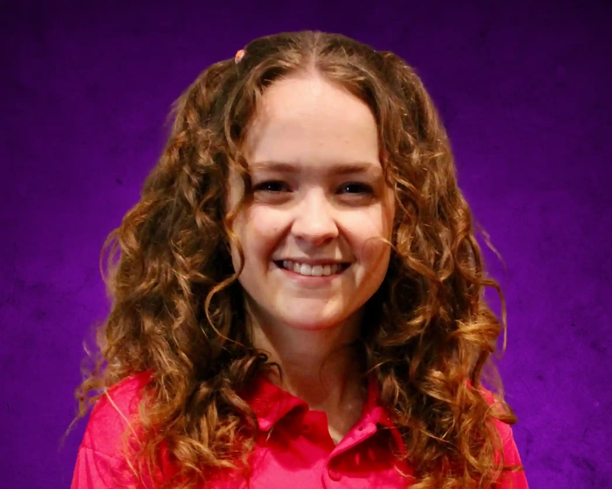 A young woman with curly hair wearing a pink shirt, smiling against a purple background.