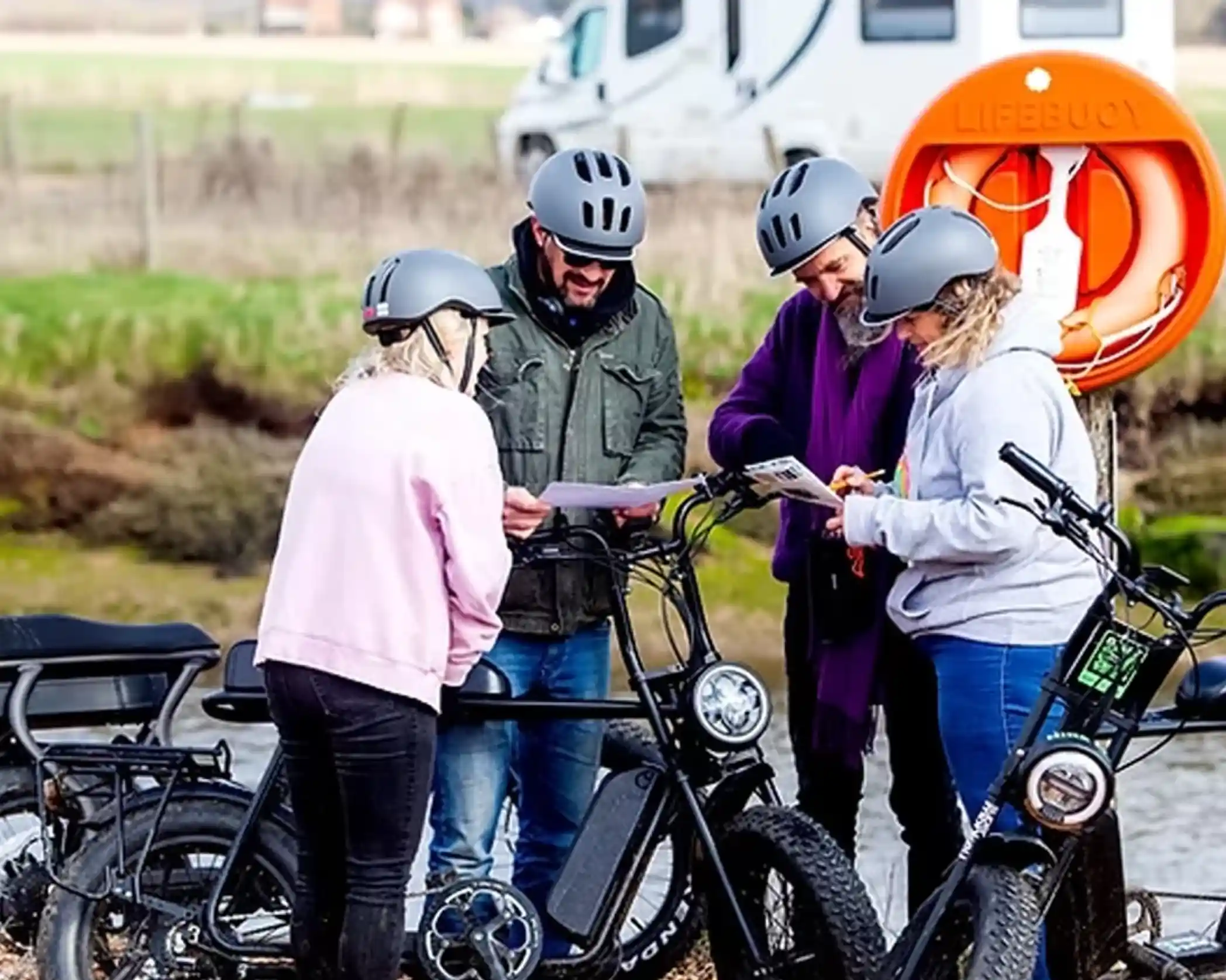 Four people wearing helmets gather around two electric bikes, studying a map. They are near a body of water with a lifebuoy prominently displayed in the background.