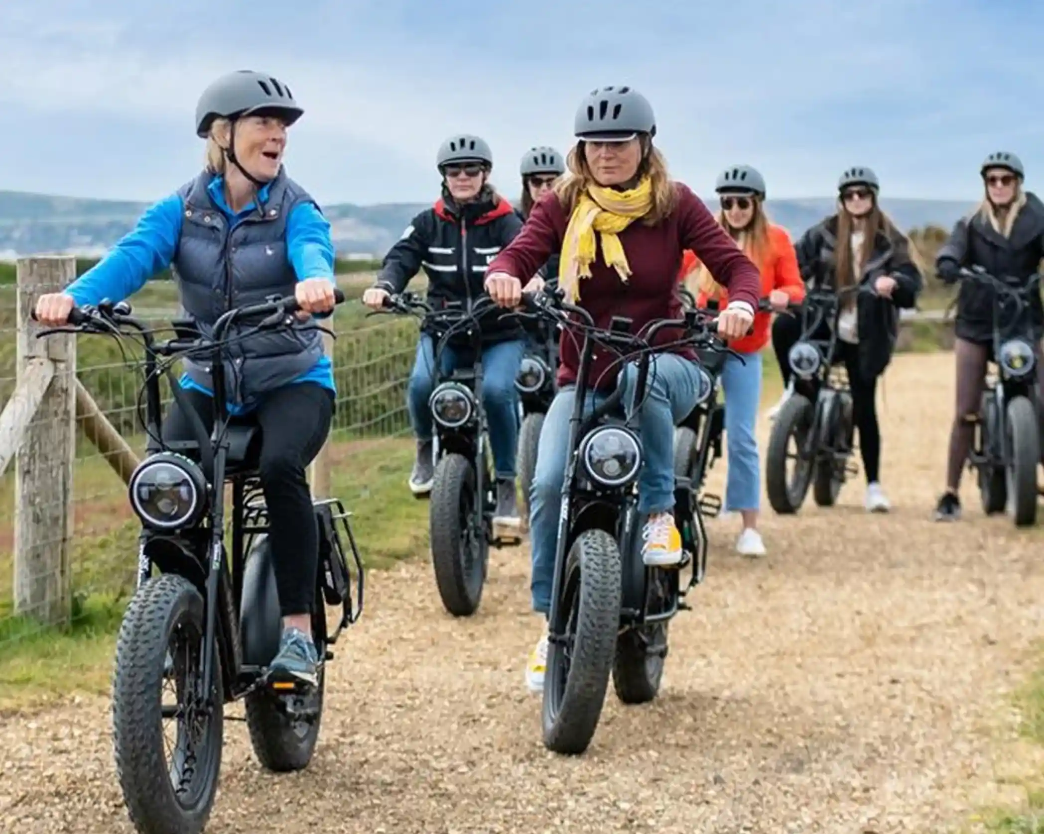 A group of seven people riding electric bikes along a dirt path, wearing helmets and casual clothing. The riders appear engaged and are enjoying a scenic outdoor environment.