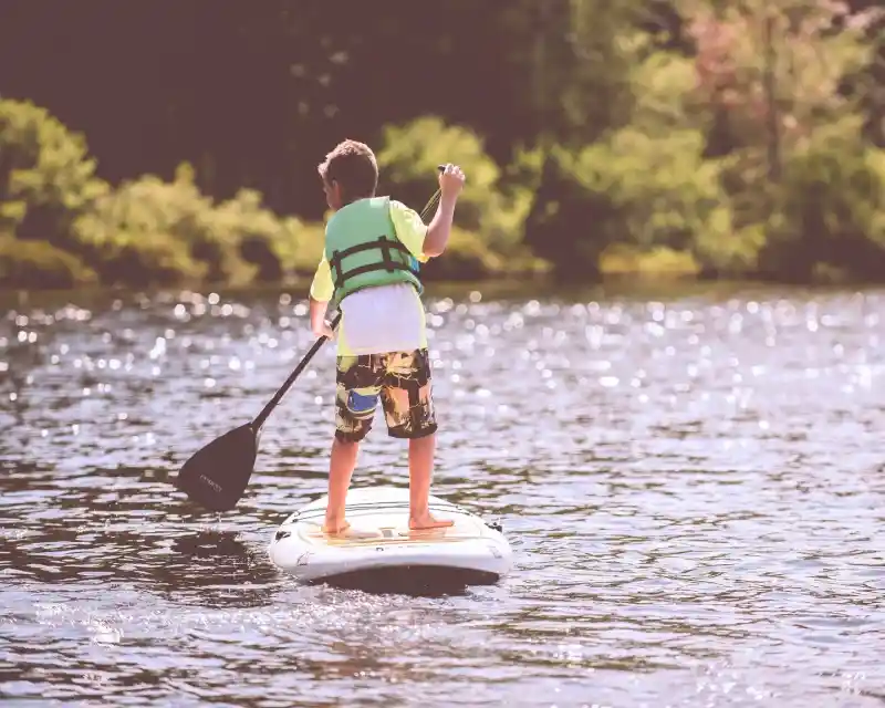 A child stands on a paddleboard, wearing a green life jacket and colorful shorts, paddling across a shimmering body of water surrounded by greenery.