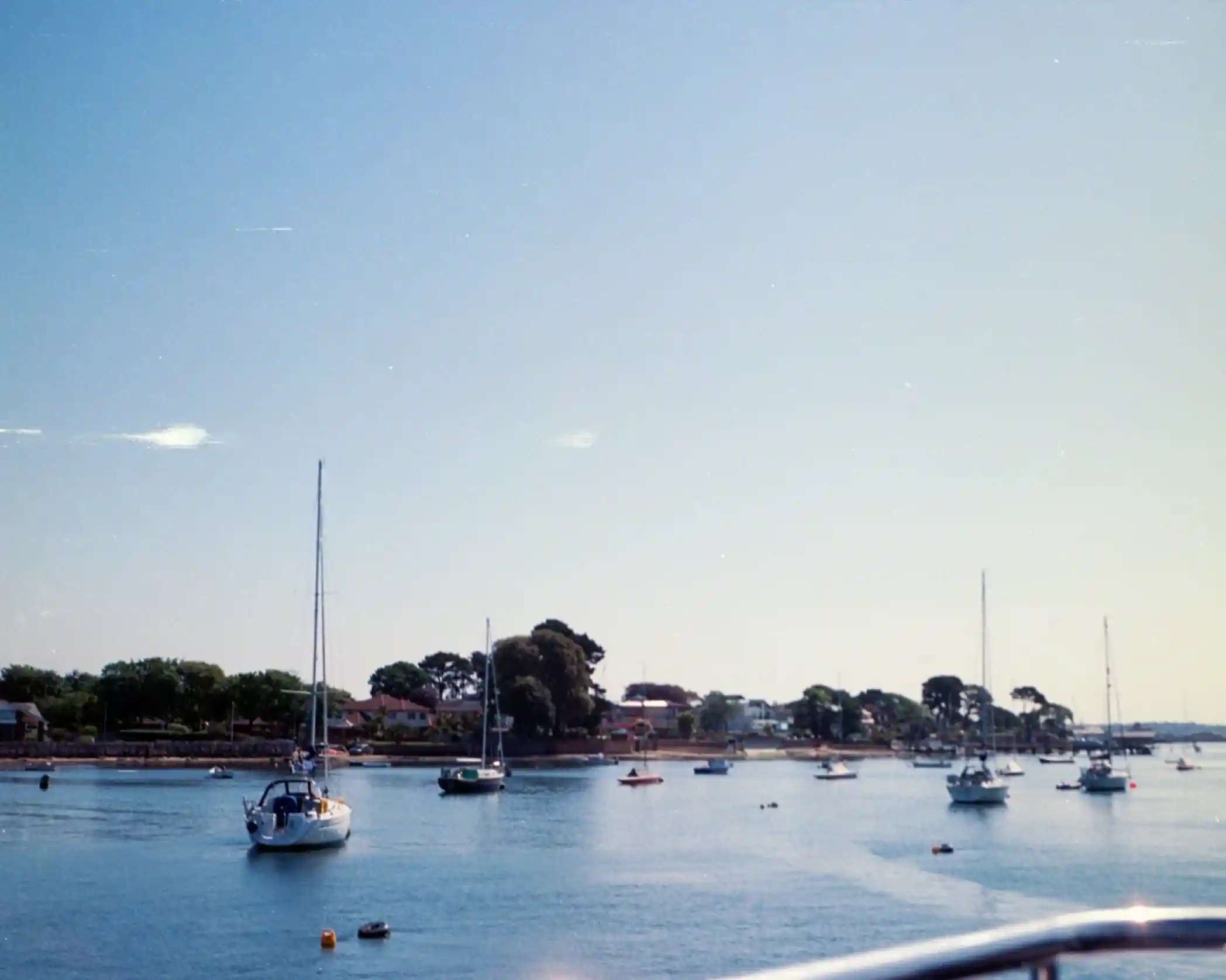 A calm harbor scene featuring several sailboats anchored in the water, with green trees and houses visible along the shoreline under a clear blue sky.