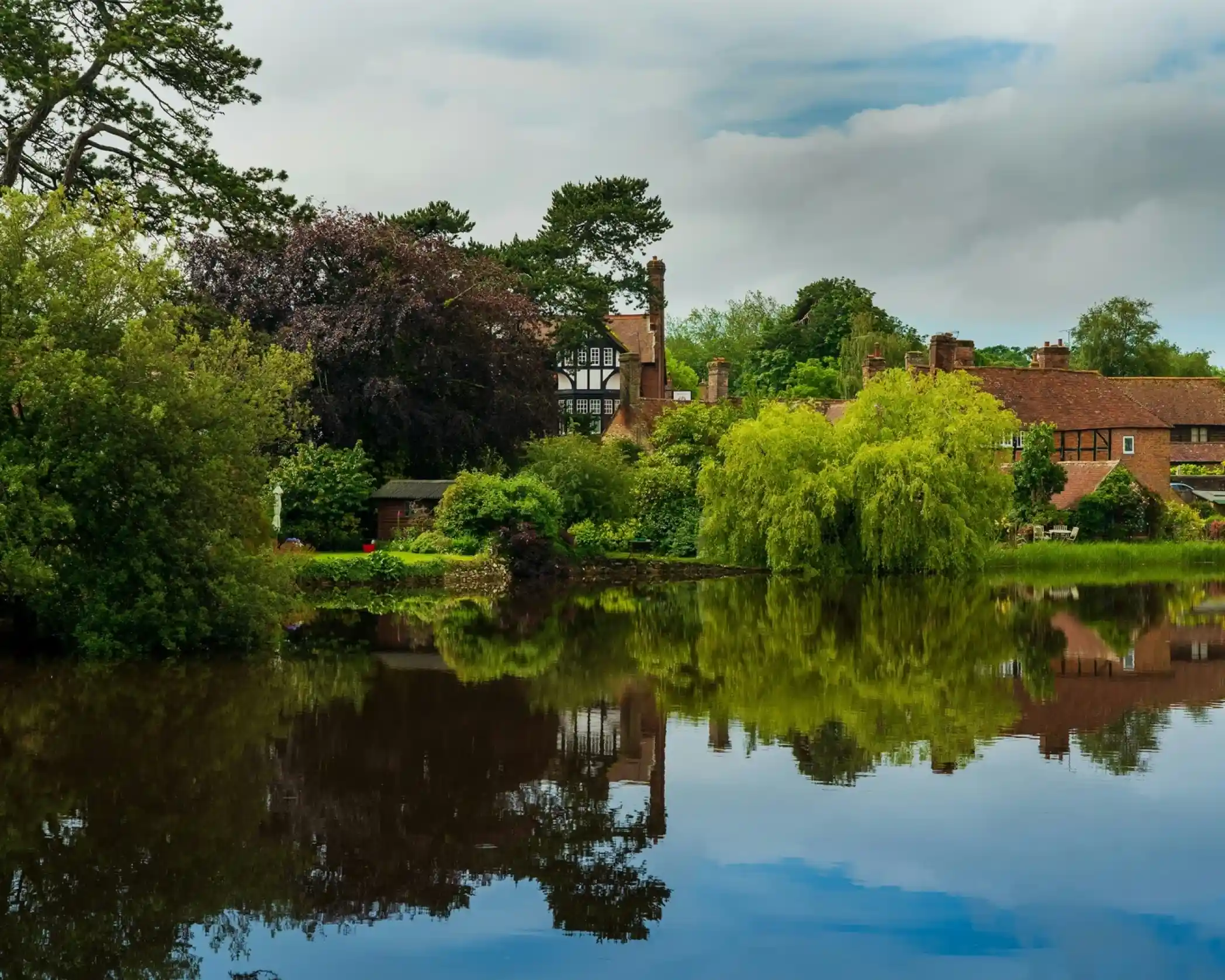 A tranquil scene featuring a serene lake reflecting lush greenery and historical architecture. Dark clouds hover above, hinting at a moody atmosphere.