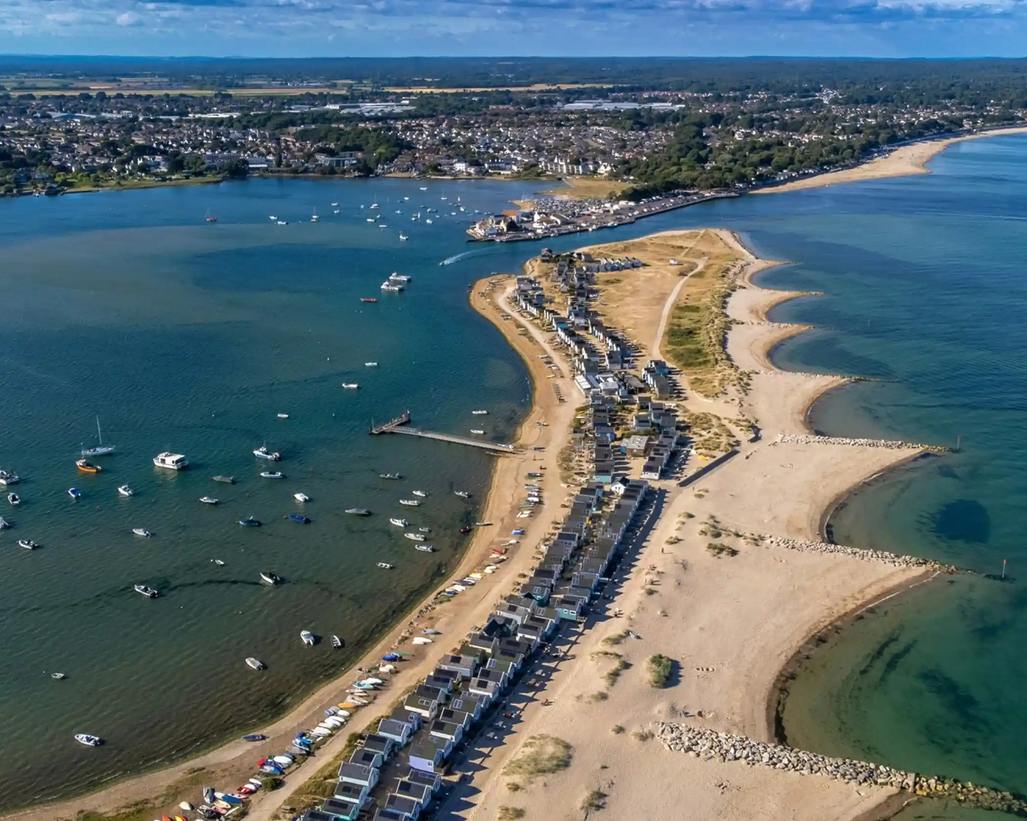 A panoramic view of a coastal area featuring sandy beaches, a marina with boats, and beachside buildings along a curving shoreline. The landscape includes a mix of water and land, with lush greenery in the background under a clear blue sky.