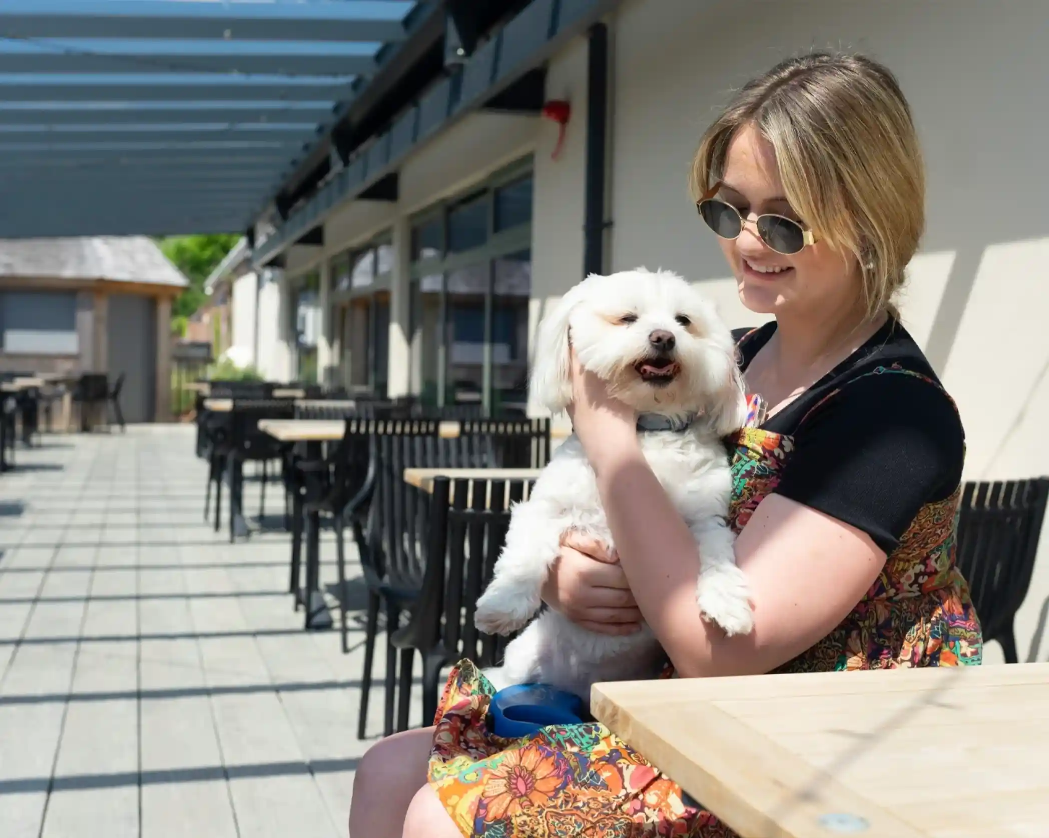 A woman wearing sunglasses and a floral dress smiles while holding a small white dog. They are seated on a patio with empty tables and a sunny atmosphere.