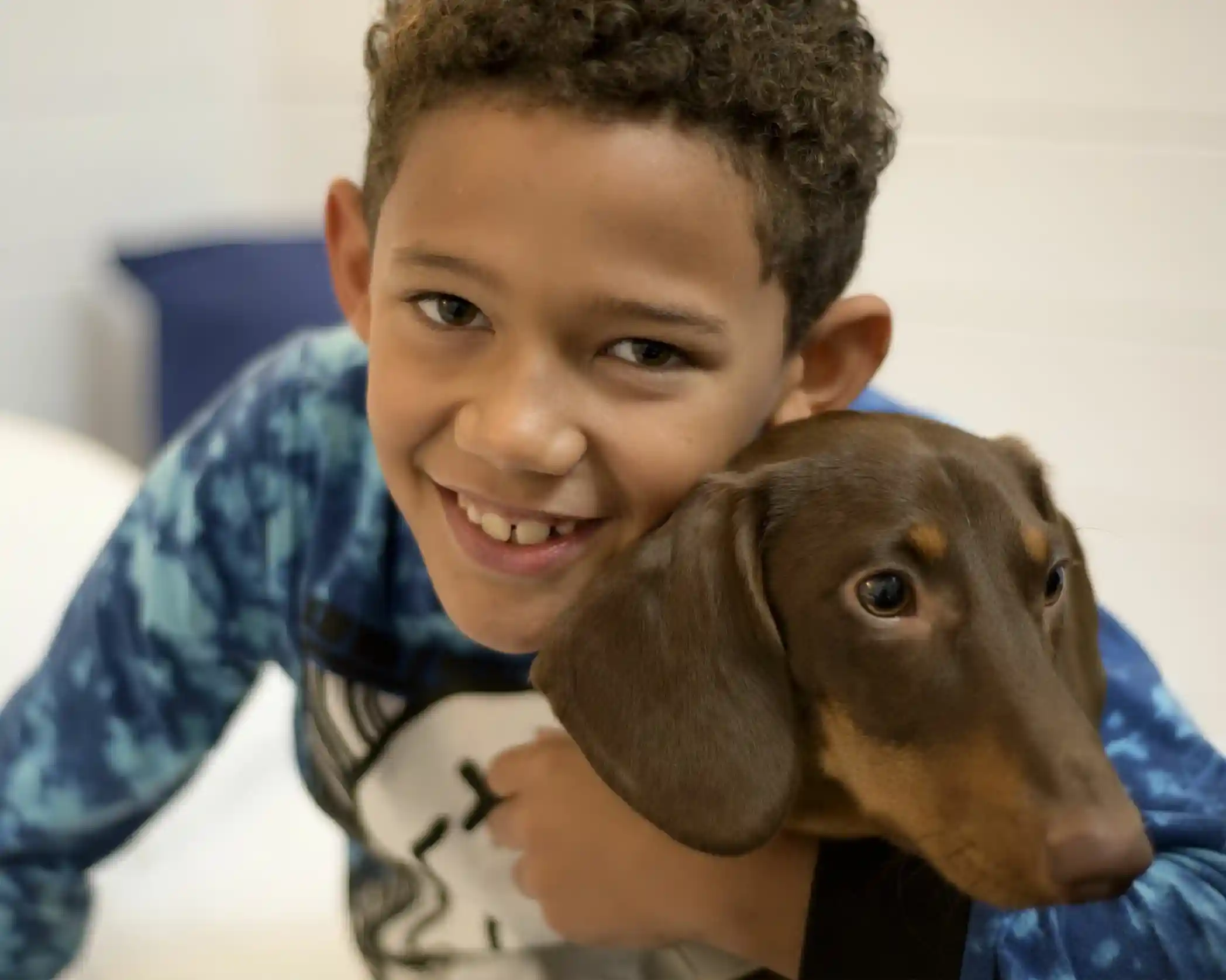 A smiling boy with curly hair hugs a brown dachshund. They both appear happy and are set against a soft, light-colored background.