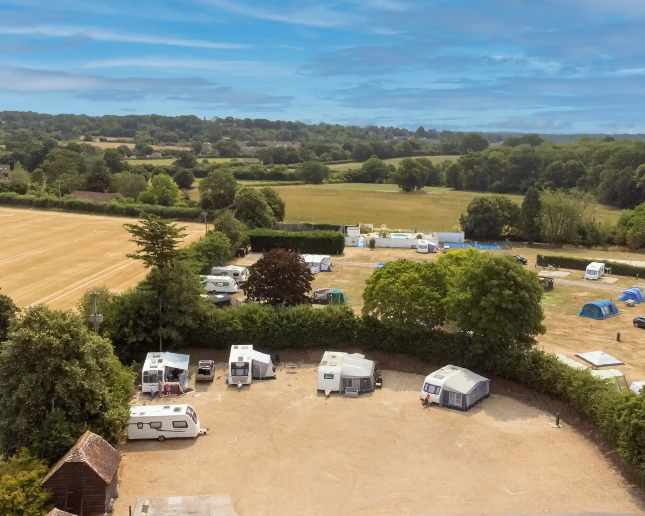 Aerial view of a caravan park with several caravans and tents set up amid lush green fields and trees. The landscape features a mix of dirt and grass pathways. The sky is partly cloudy, adding a serene atmosphere to the scene.